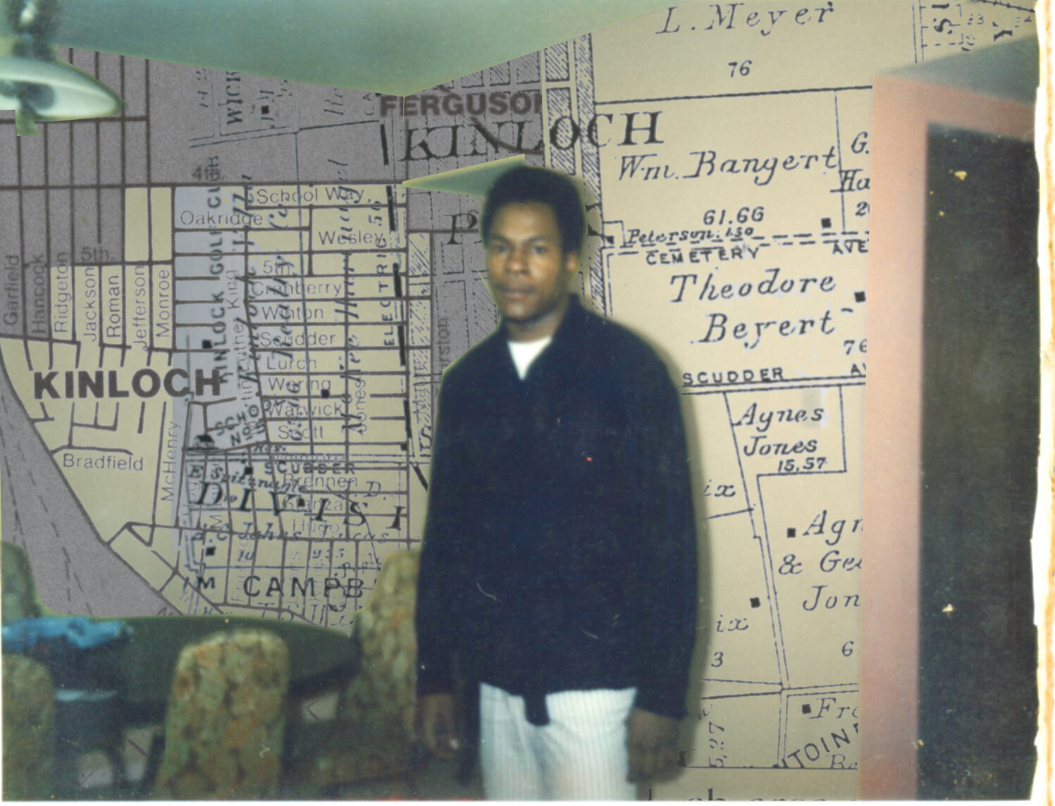 An aged color photo of an adult man with medium dark skin and short dark hair looks forward with a long sleeve dark blue shirt and white pinstripe pants, standing in front of a wallpapered surface meant to resemble an antique neighborhood map that combines various plot grids, and different font texts featuring words like: “Kinloch,” and “Ferguson.”
