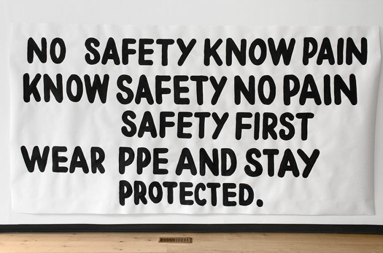 A large piece of paper mounted on a wall, with words painted onto it. The words read: "No Safety Know Pain. Know Safety No Pain. Safety First. Wear PPE and Stay Protected."