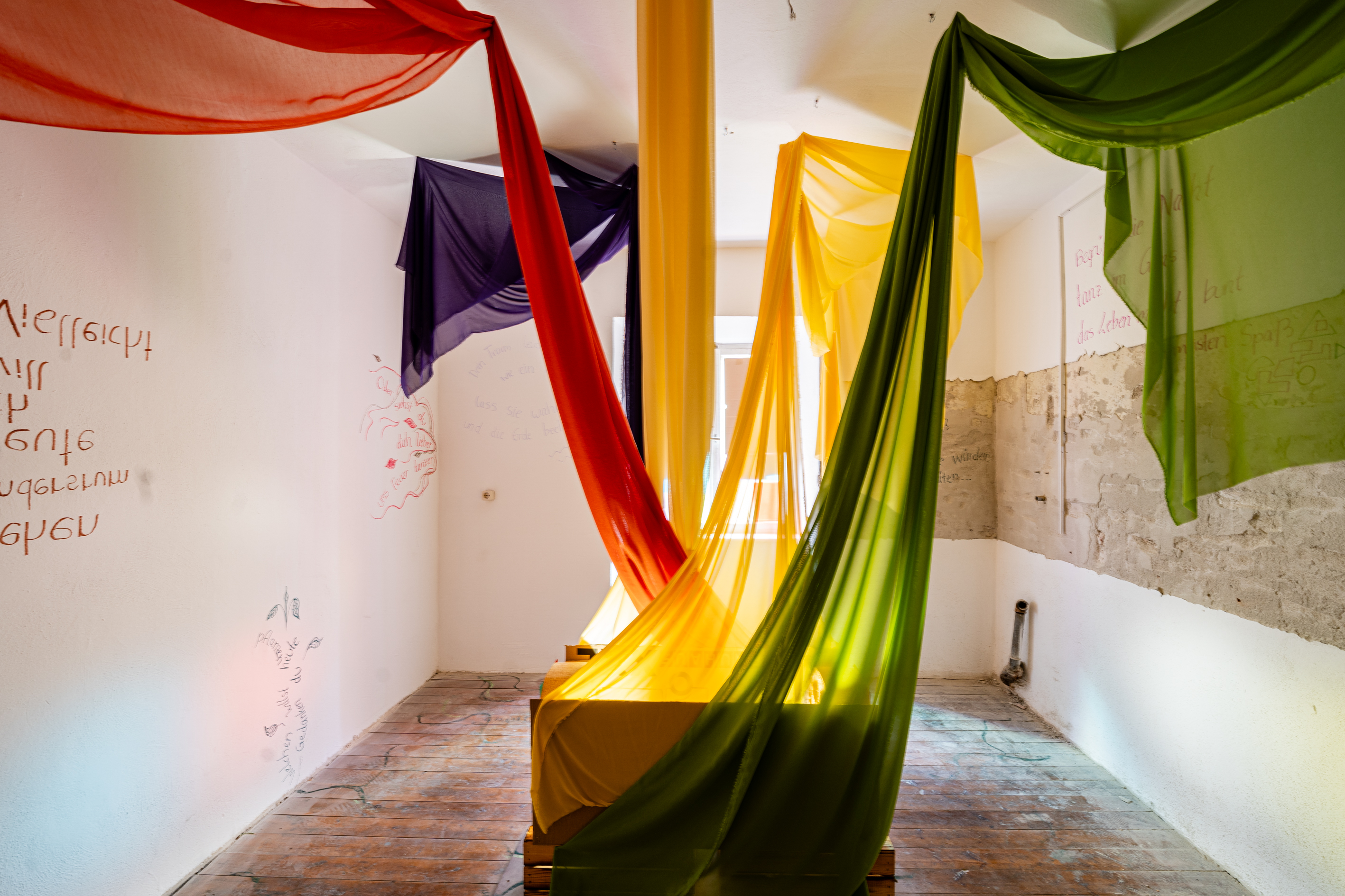 Bild 1 Poetree: Ein wei&szlig;er Raum, mit T&uuml;chern in Gr&uuml;n, Gelb , Lila und Rot, die in unterschiedliche Richtungen an der Decke aufgeh&auml;ngt wurden. Sie &uuml;berschneiden sich alle in der Mitte des Raumes.