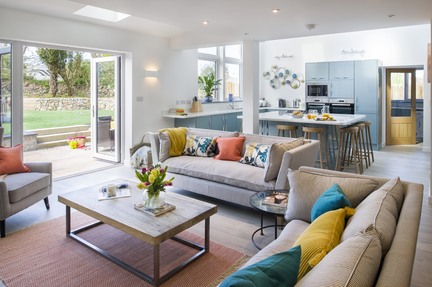 Light blue kitchen with breakfast bar and wooden stools. Lounge with beige sofas and orange, yellow, teal soft furnishings.