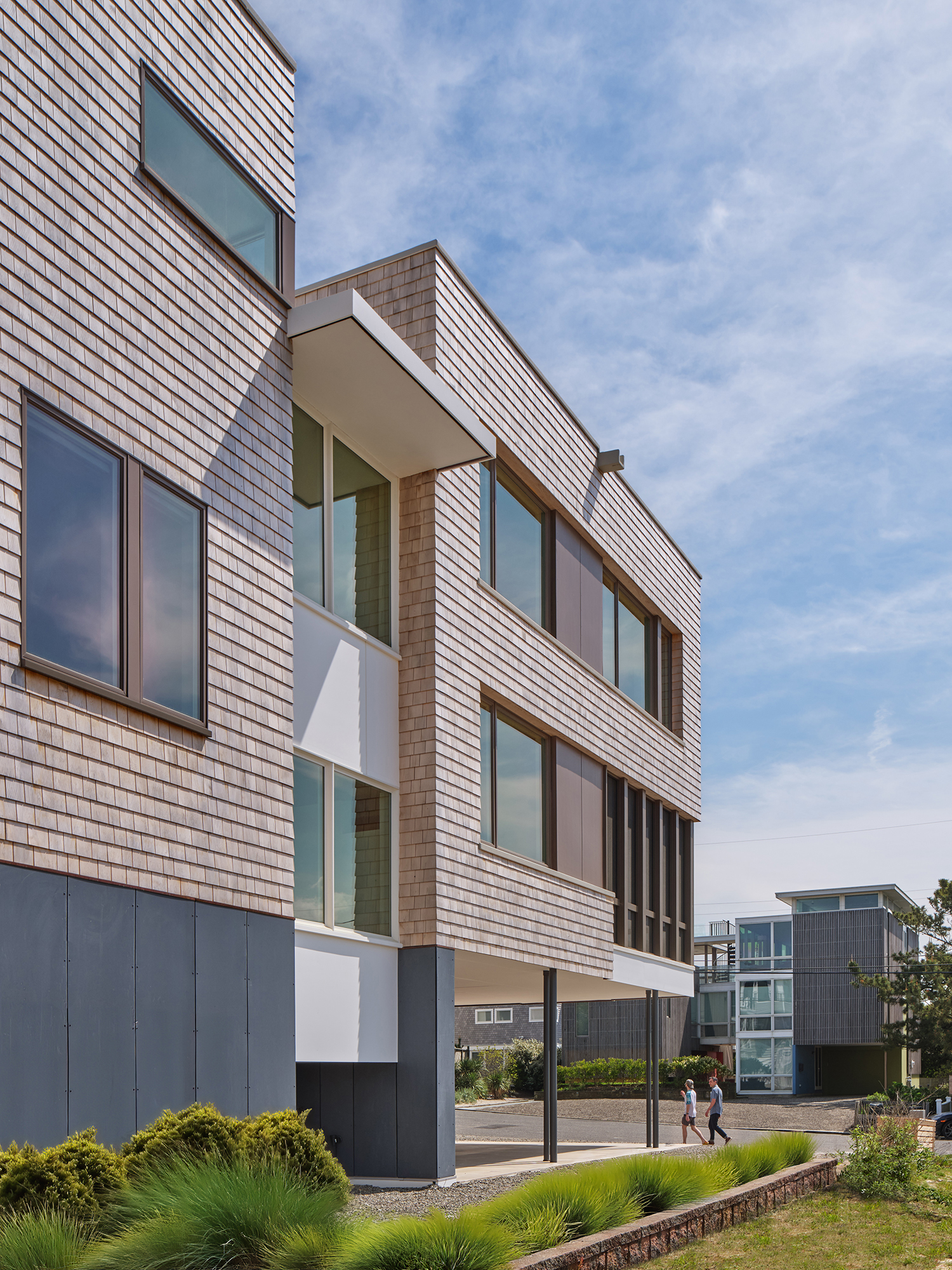 Exterior view of the Gap House West Elevation from a low-angle perspective, showcasing the modern shingle cladding, varied window placement, and the elevated structure with thin columns.