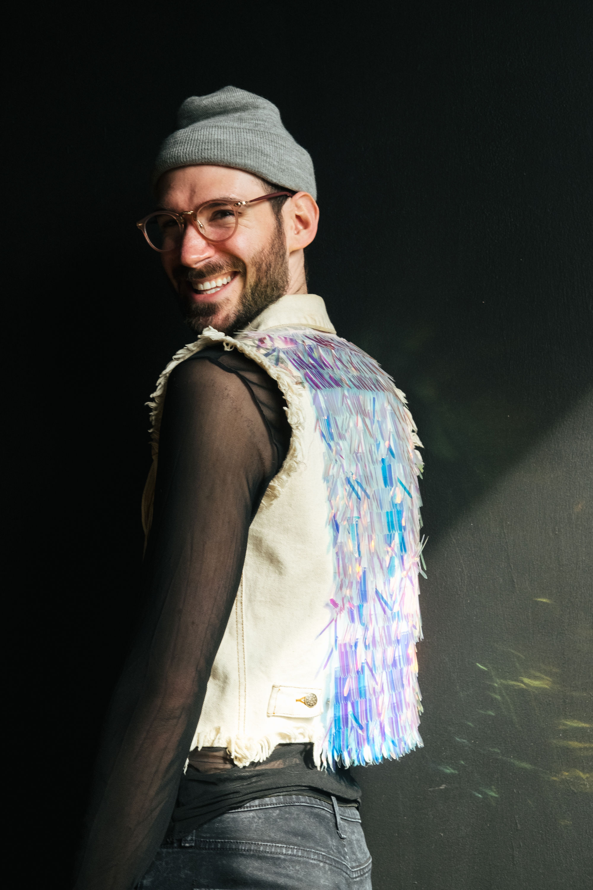 Image description: Kevin Gotkin faces away from the camera and smiles over their shoulder. They wear glasses, a gray beanie, and a sheer black long-sleeve with a white vest over it. The white vest has an iridescent design on the back that shines in the sunlight.