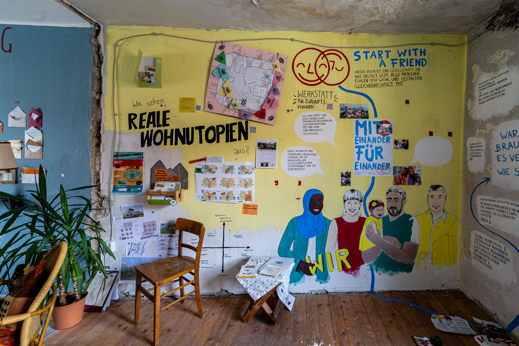 Bild 1 zu Reale Wohnutopien: Eine gelbe Wand beh&auml;ngt mit Informationen zum Wohnprojekt Sherlo. Dar&uuml;ber steht in schwarzen Druckbuchstaben: Wie sehen reale Wohnutopien aus? Der rechte Teil geh&ouml;rt zu Start with a friend.  Links vor der Wand steht eine Pflanze und ein Holzstuhl.