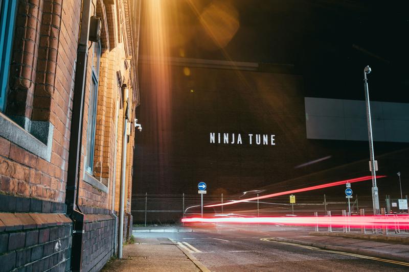 NINJA TUNE text projection displayed on a brick wall in Belfast at night with light trails crossing the frame.
