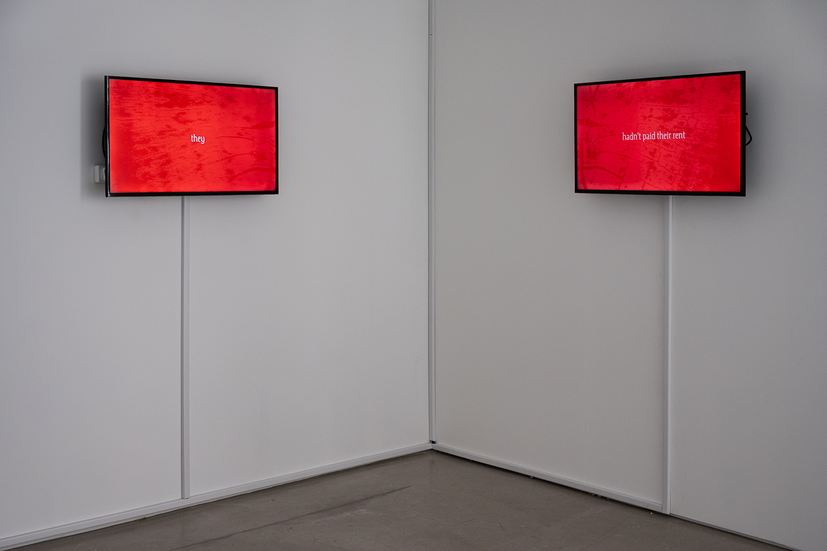 The image shows two flat screen monitors mounted perpendicular to one another, at the corner of two white gallery walls. Both monitors display white gothic text on a textured red background. On the left hand screen the text reads 'They' and on the right hand screen the text reads 'hadn't paid their rent.'