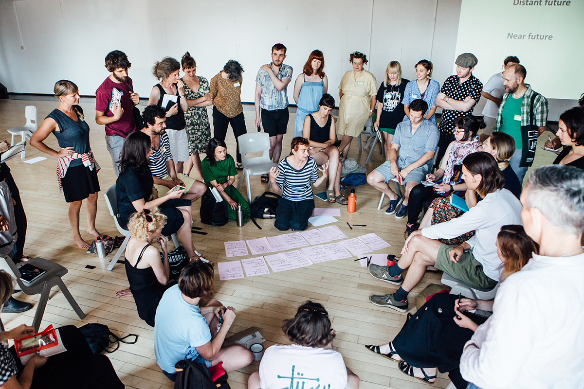A group of Jamboree participants stand in a circle looking at notes spread over the floor while the leader of the group speaks.