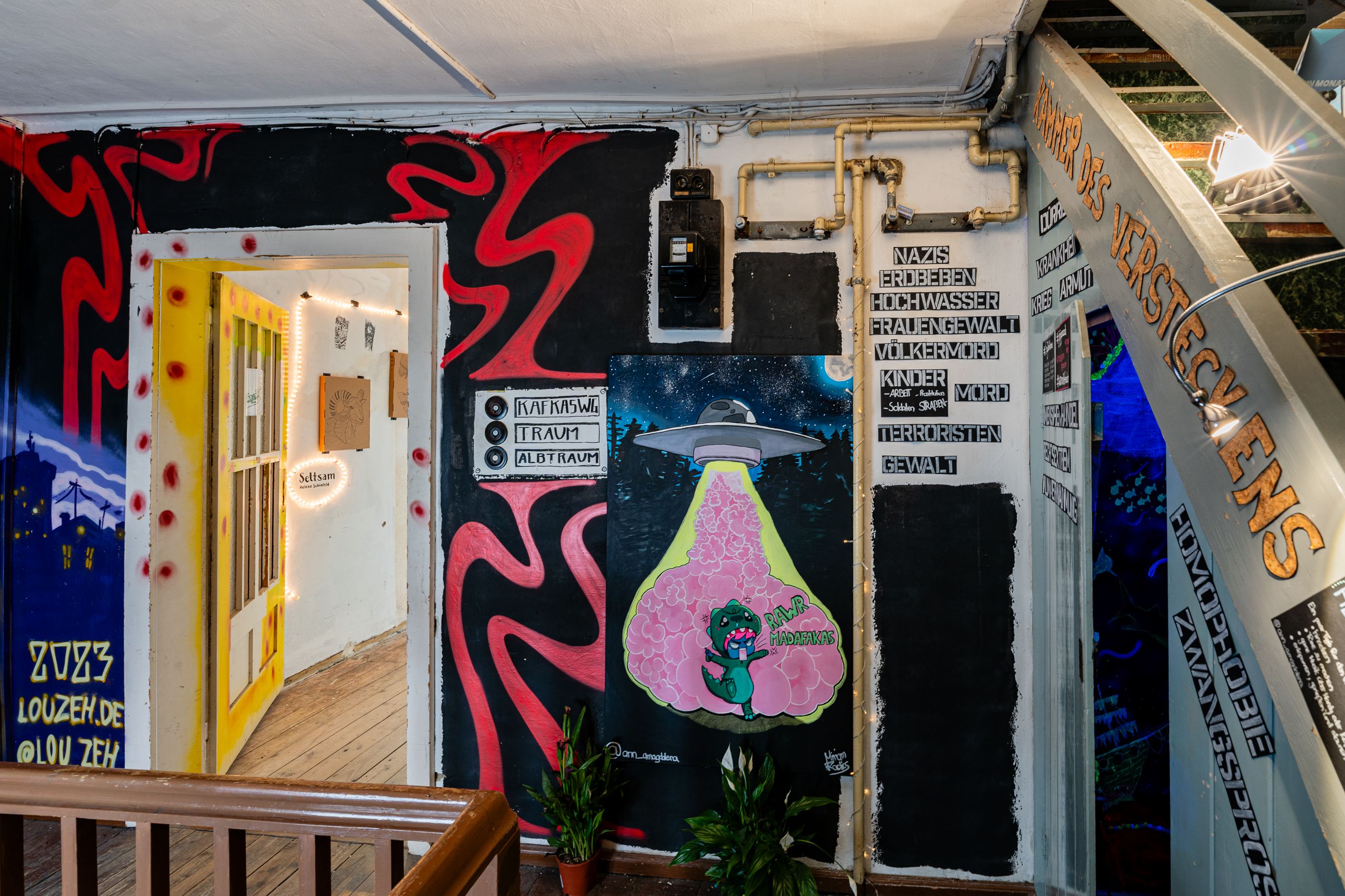 Bild 3 zu Beyond Wildest Dreams: Die Wand um den Eingang zu Kafkas WG ist dunkel mit roten geschlungenen Formen bemalt. Rechts davon erkennt man ein Ufo. Wasserrohre sind golden bemalt und die Kammer des Versteckens befindet sich rechts unterhalb der Treppe zum Dachboden. 