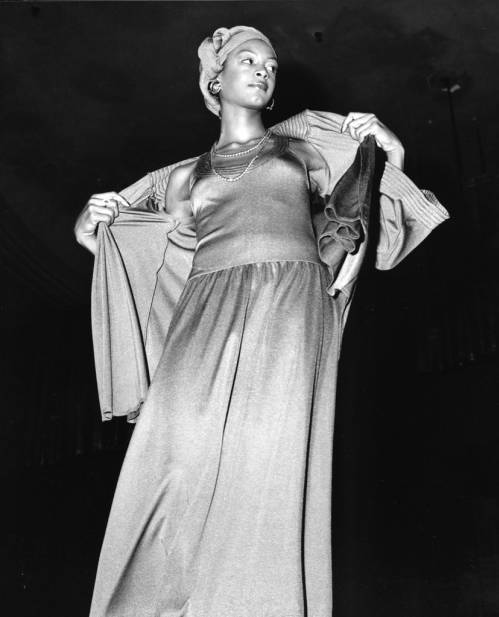 Image: A black-and-white photo of an Ebony Fashion Fair model posing on a runway in a long dress and jacket, pulling it open, c. 1974. She’s looking off to the right, wearing a head wrap and earrings. African American Photographs Collection, Tom and Ethel Bradley Center, California State University Northridge. Photographer: Guy Crowder.