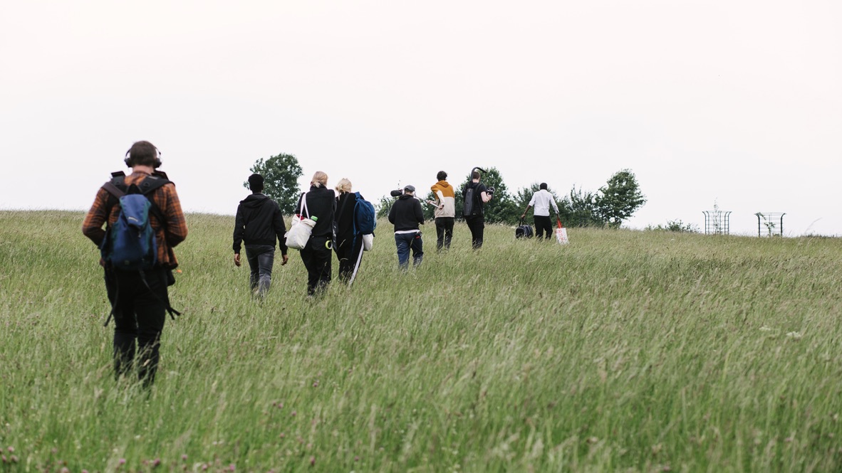 The crew of Son of Helios follow the cameraperson in a line through a grassy meadow while two young performers storm ahead during a take. The grass is waist-high.