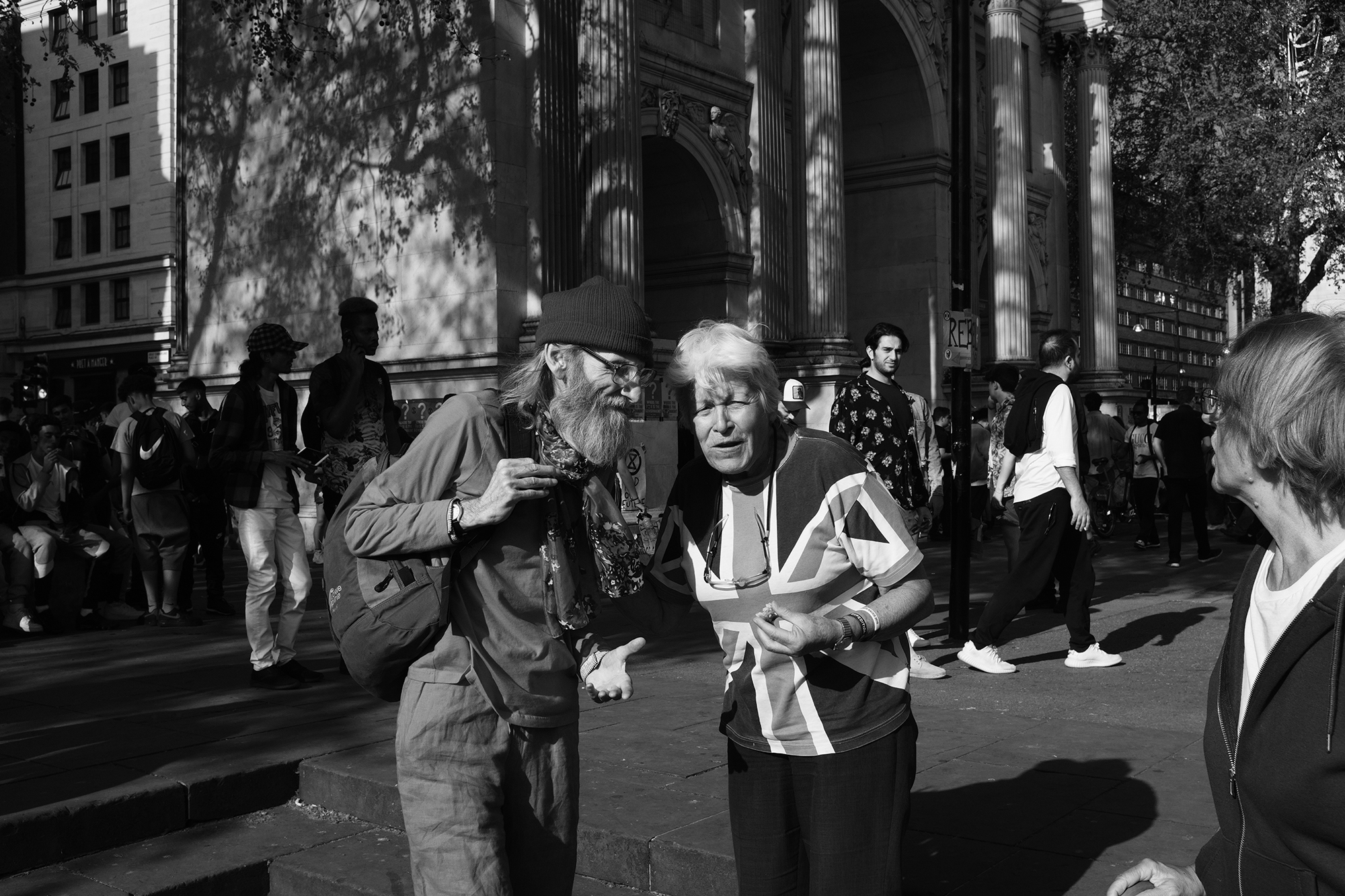 Philippe-Sarfati-reportage-photography-photographer-street-documentary-photojournalism-photojournalist-london-uk-xr-extinction-rebellion-black-and-white-old-man-woman-explaining-british-flag-shirt-marble-arch-gathering
