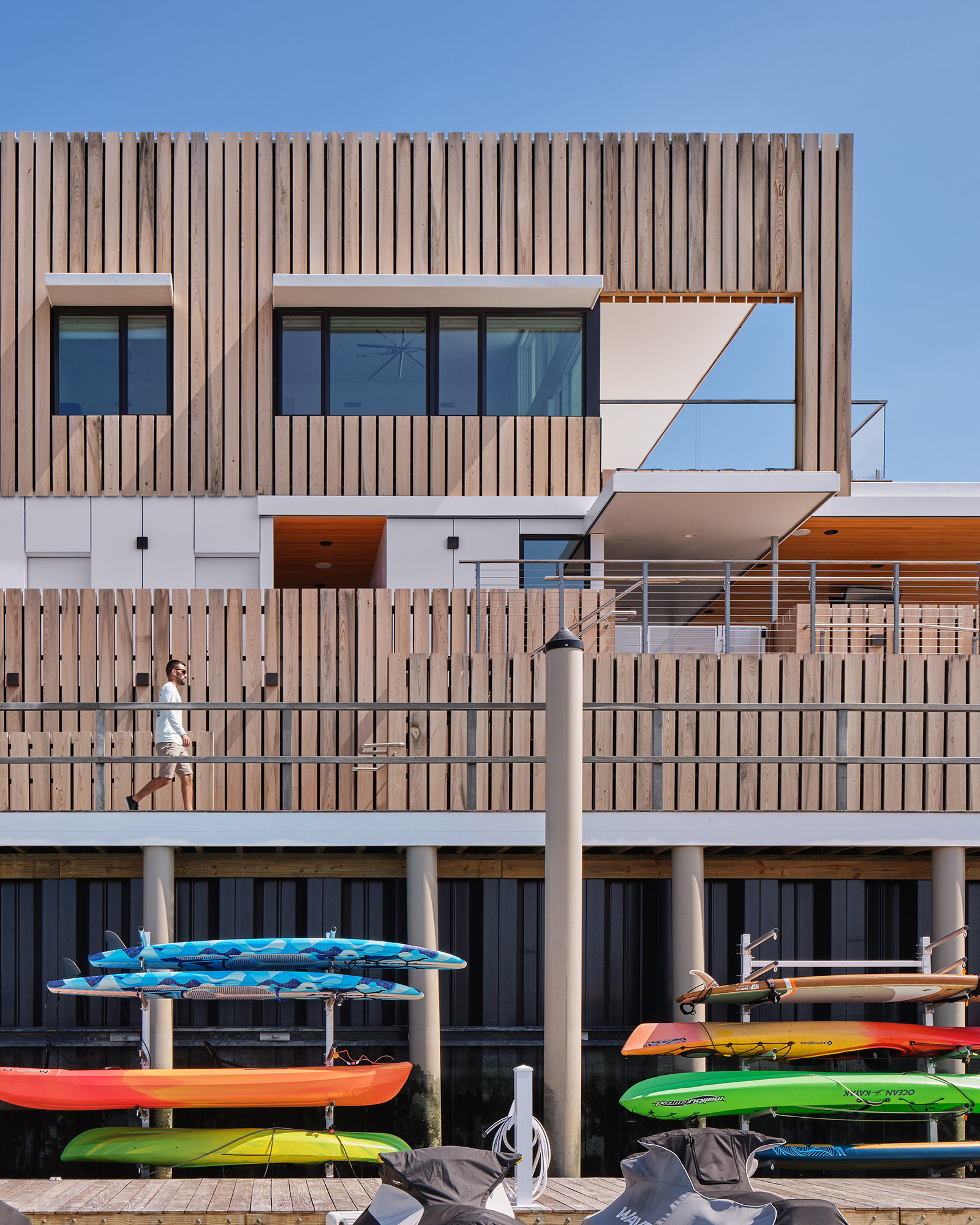 Exterior view of the Brigantine Paddle Club's waterside facade, showing colorful kayaks and paddleboards stored beneath the elevated deck and the modern vertical Cypress rainscreen cladding above.