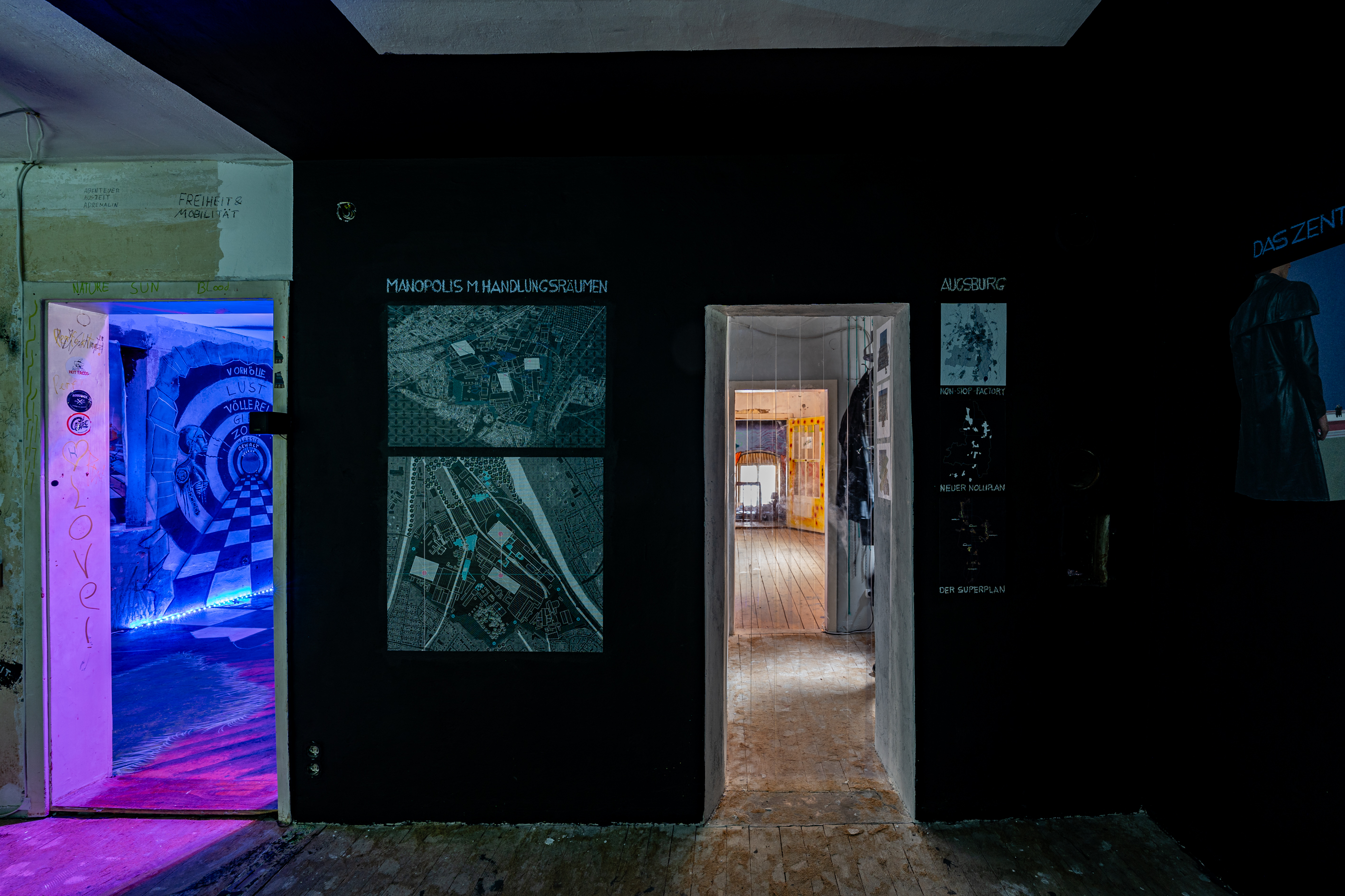 Bild 5 zu Die Welt als Fabrik: Blick auf eine Wand und einen Durchgang zum Flur. Links des T&uuml;rrahmens befinden sich zwei  Stadtpl&auml;ne, rechts der T&uuml;r ebenfalls drei. Davon ist der oberste von Augsburg.