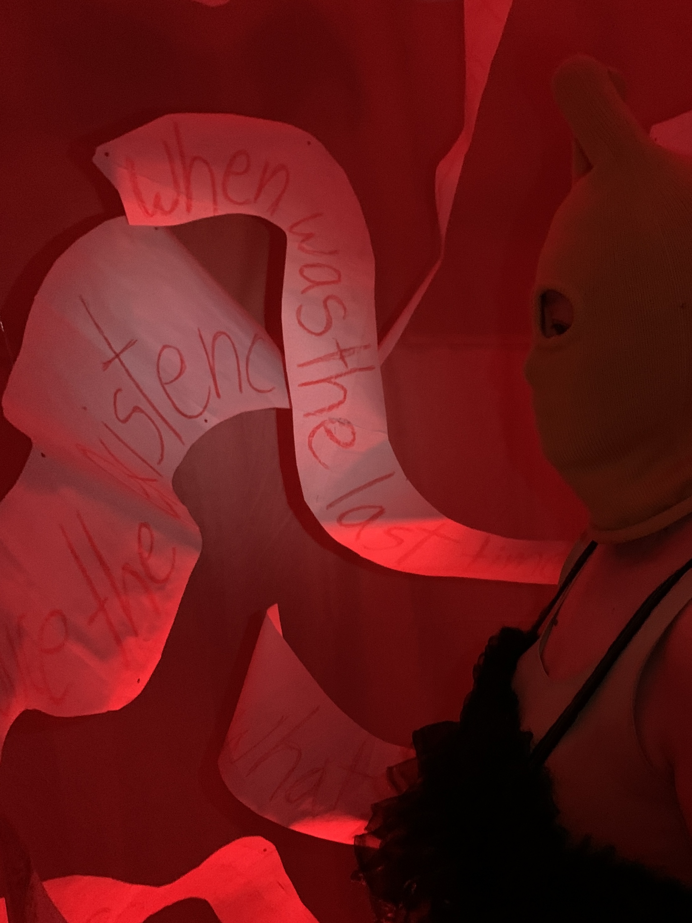 Another view of a person in a mask observing the words that are made seen in the red with a white flashlight pointed towards the suspended paper sculpture