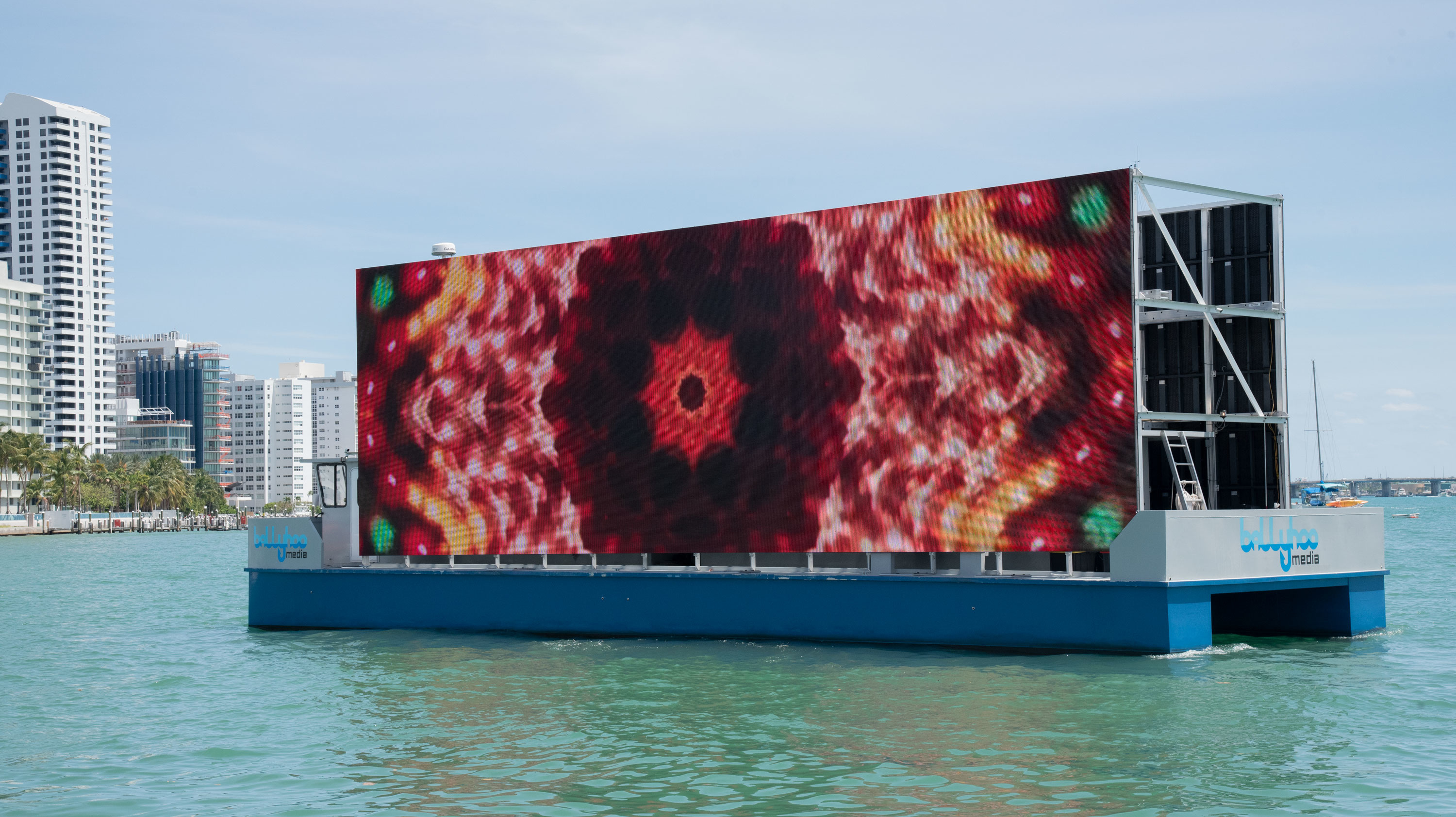 Displayed on a large screen that is being exhibited by boat, Carlos Betancourt’s 2020 video installation projects a primarily red kaleidoscope image that has yellow and green accents.