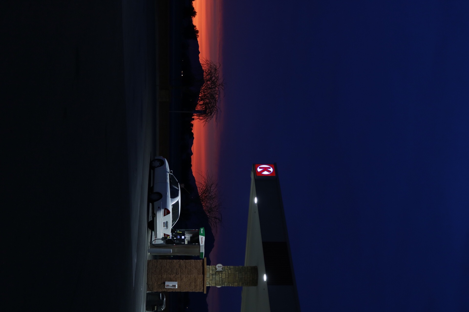 A white car is parked at a dimly lit gas station under a dark sky. In the background, a vibrant orange sunset silhouetted by mountains creates a serene atmosphere, in Phoenix, AZ.