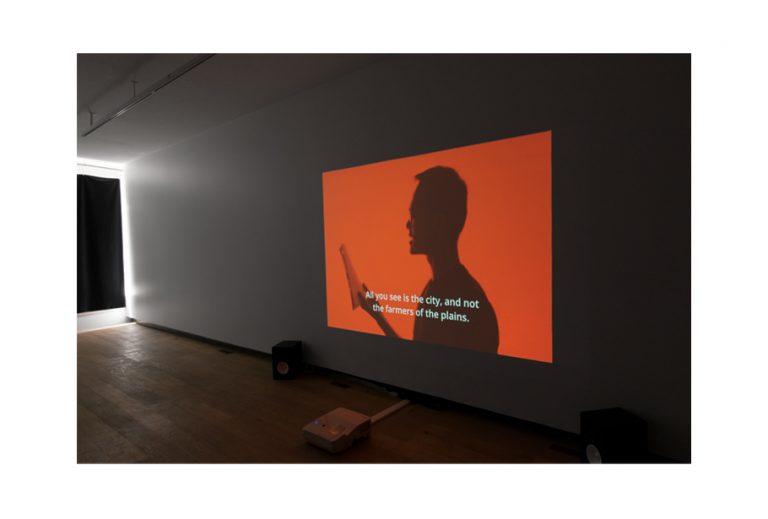 A photo of the projected movie "The Young Comrade" on a white wall in the gallery. The still shot in the movie is a silhouette of a man with glasses on a red background. The man is holding and reading a book. The subtitle reads: "All you see is the city, and not the farmers of the plains."  