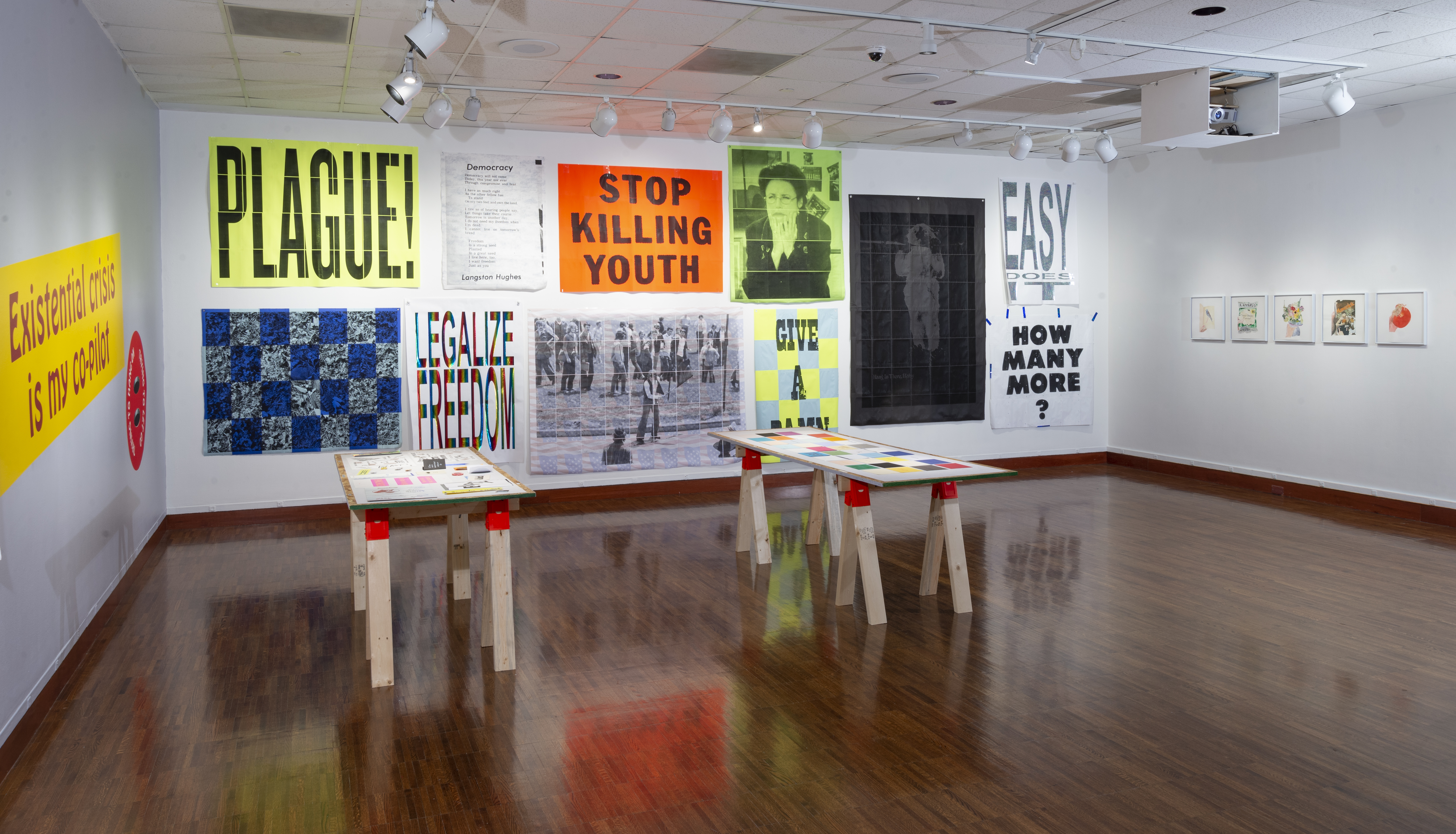 A white walled gallery with two sawhorse tables in the center with small pieces of paper works on top, and lined all around, and completely covering the walls, are various posters or different shapes and colors that read things like “Plague!” or, “Stop Killing Youth!”