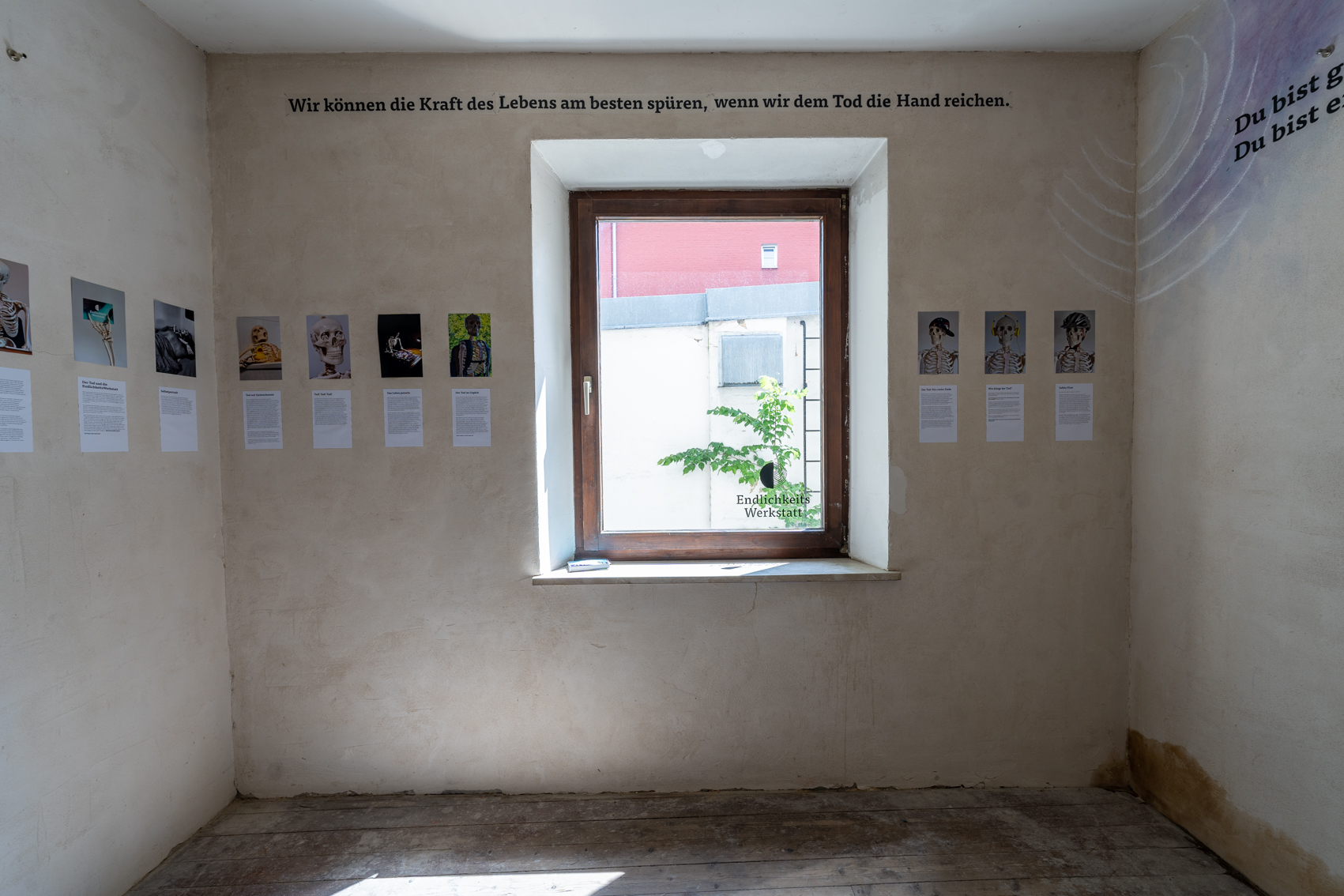 Bild 2 zu Augsburger Totentanz: An der Wand mit dem Fenster gegen&uuml;ber des Eingangs, h&auml;ngen auf Augenh&ouml;he Bilder von Skeletten bei menschlichen T&auml;tigkeiten, wie zum Beispiel mit Kopfh&ouml;rern, Fahrradhelm oder in der Badewanne. Darunter befinden sich Texte. &Uuml;ber dem Fenster steht an der Wand: "Wir k&ouml;nnen die Kraft des Lebens am besten sp&uuml;ren, wenn wir dem Tod die Hand reichen."