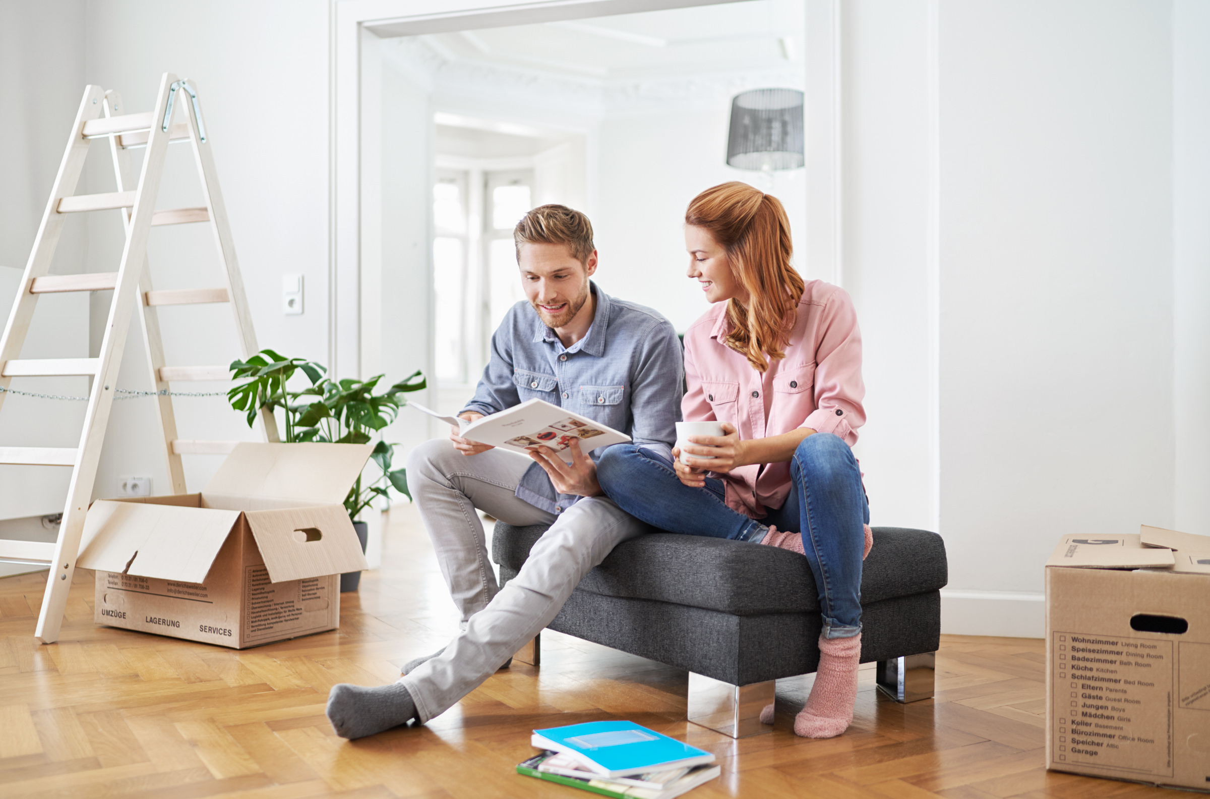 Portrait eines Paerchens, sitzend auf einem Couchhocker. Umzugskisten deuten auf einen Umzug hin.