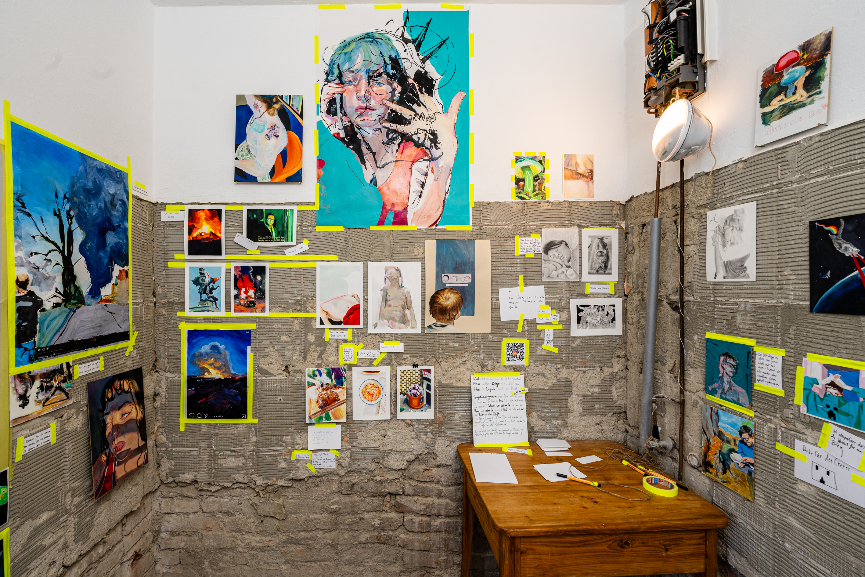 Bild 3 zu Kunst kommt von Kompostieren: Wand gegen&uuml;ber des Eingangs. Zeigt ebenfalls Malereien. Ein Holztisch mit Stift, Zettel und Klebeband steht in der rechten Ecke des Raumes.