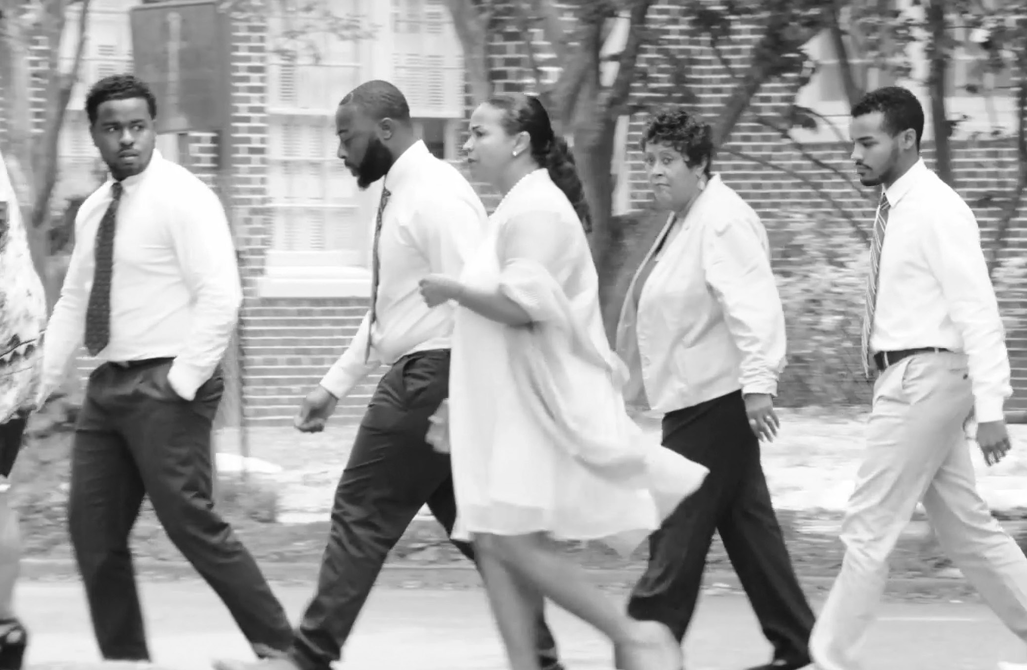 A group of five Black people, all dressed in white shirts, moves to the left of the screen. One person is slightly in the foreground, wearing a flowy white dress, while the four people walking in the background all wear pants (the first three have blank pants, the last one is in khakis). The person at the very front of this group is looking back, at the person in the foreground, who looks on. This is the same photo as right above, only slightly zoomed in.