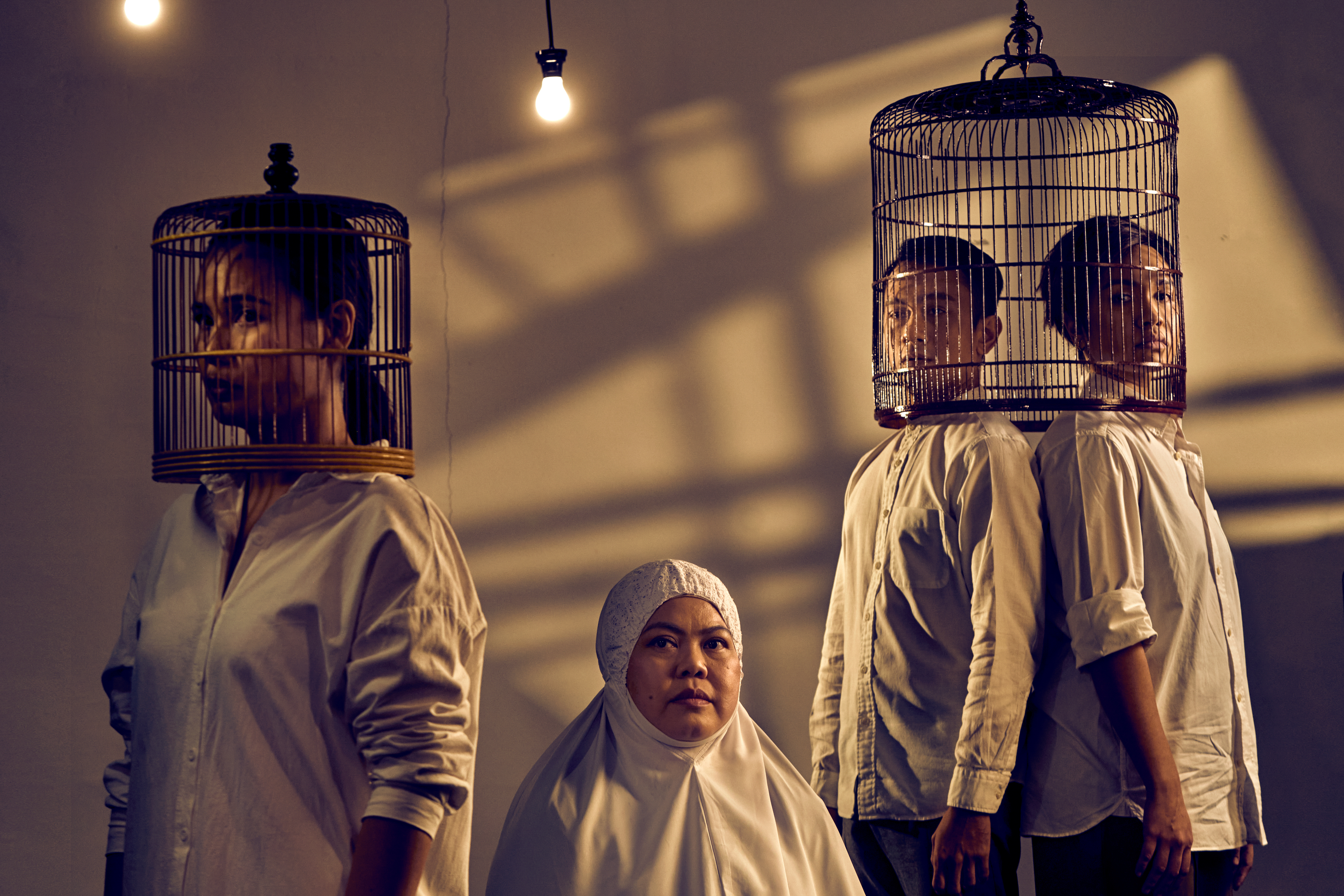 Image 1 of 5: Three persons wearing bird cages over their heads, and a lady in a Malay prayer garment