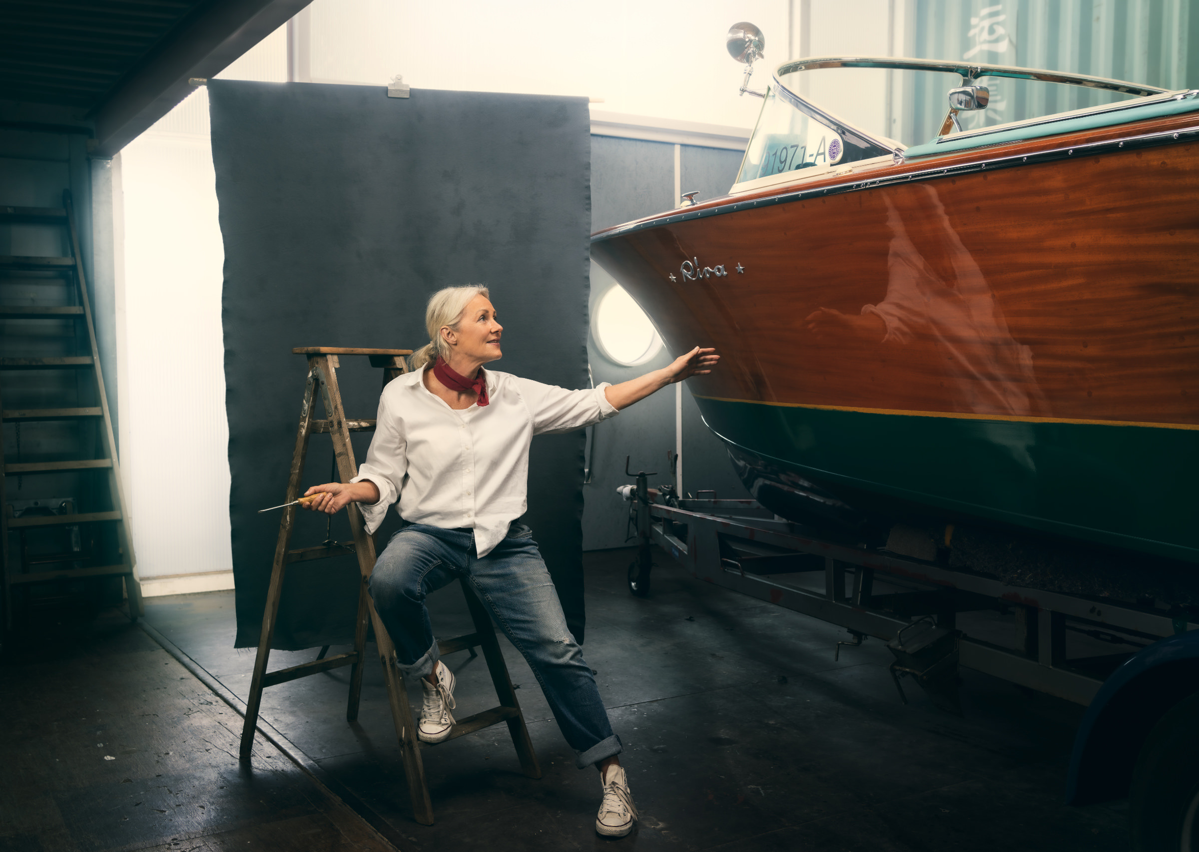 Environmental Portrait aus der Serie The Workshop mit einem alten Riva-Boot. In einer Bootswerkstatt sieht man eine aeltere blonde Frau in Jeans und weißer Bluse sitzend auf einer Leiter  vor einem Stoffhintergrund neben einem alten Riva Holzboot.