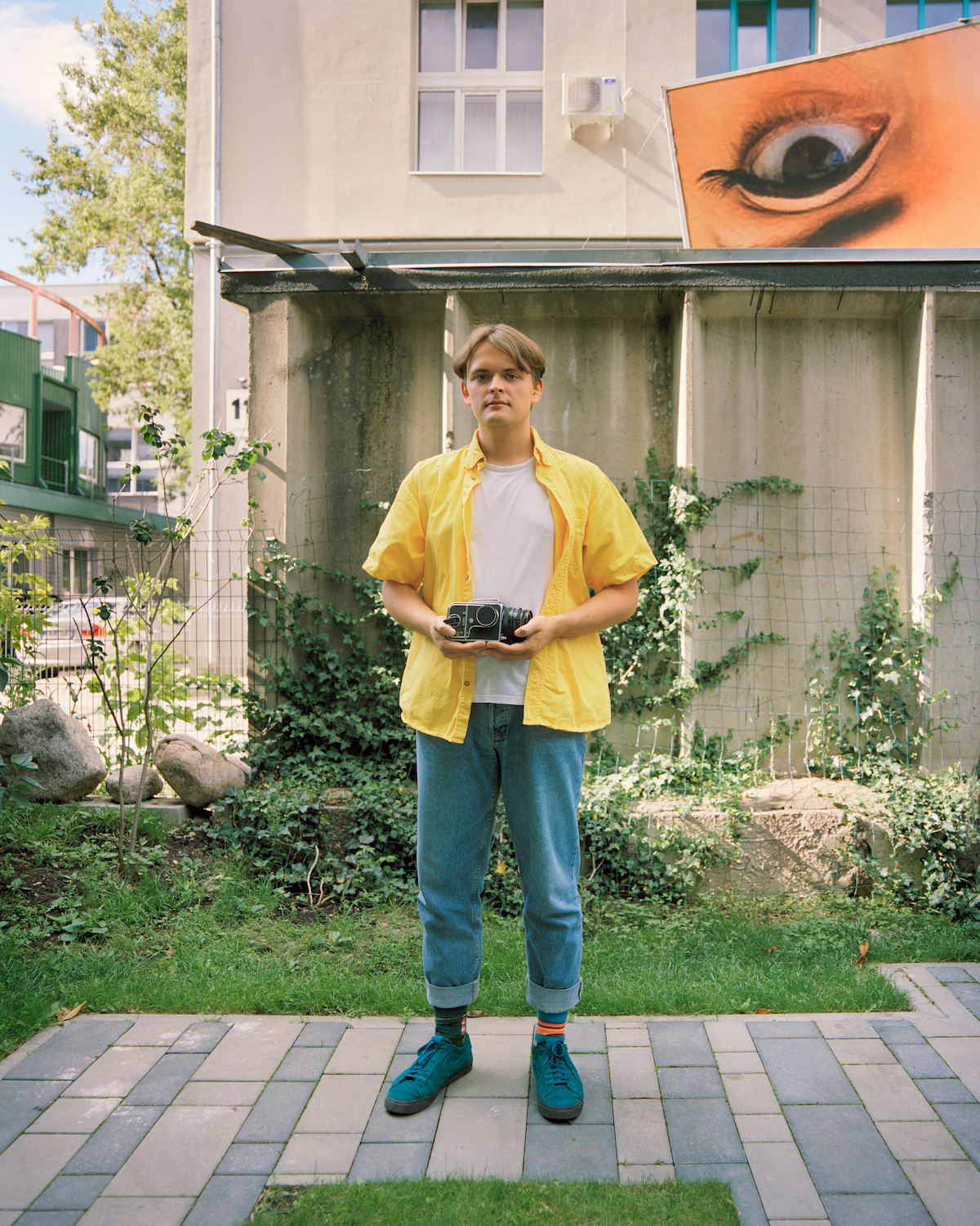 Jonas Balsevičius, g. 2001 m. Jonas gimė ir užaugo Vilniuje. Vizualiuosiais menais vaikiną paskatino domėtis dėdė, kuris yra fotografas ir videografas, padovanojęs pirmąjį Jono juostinį fotoaparatą “Hasselblad”. Dabar vaikinas užsiima meno renginių kuravimu bei fotografija. Jo portretas buvo įamžintas Vilniuje Ševčenkos gatvės loftų vidiniame kieme, kuriame jis pradėjo domėtis fotografija ir kava. Ši eklektiška lokacija reprezentuoja Jono tarpdisciplinišką kūrybą, kurios pagrindas yra fotografija. Dabar vaikinas studijuoja meninę fotografiją Londono menų universitete.