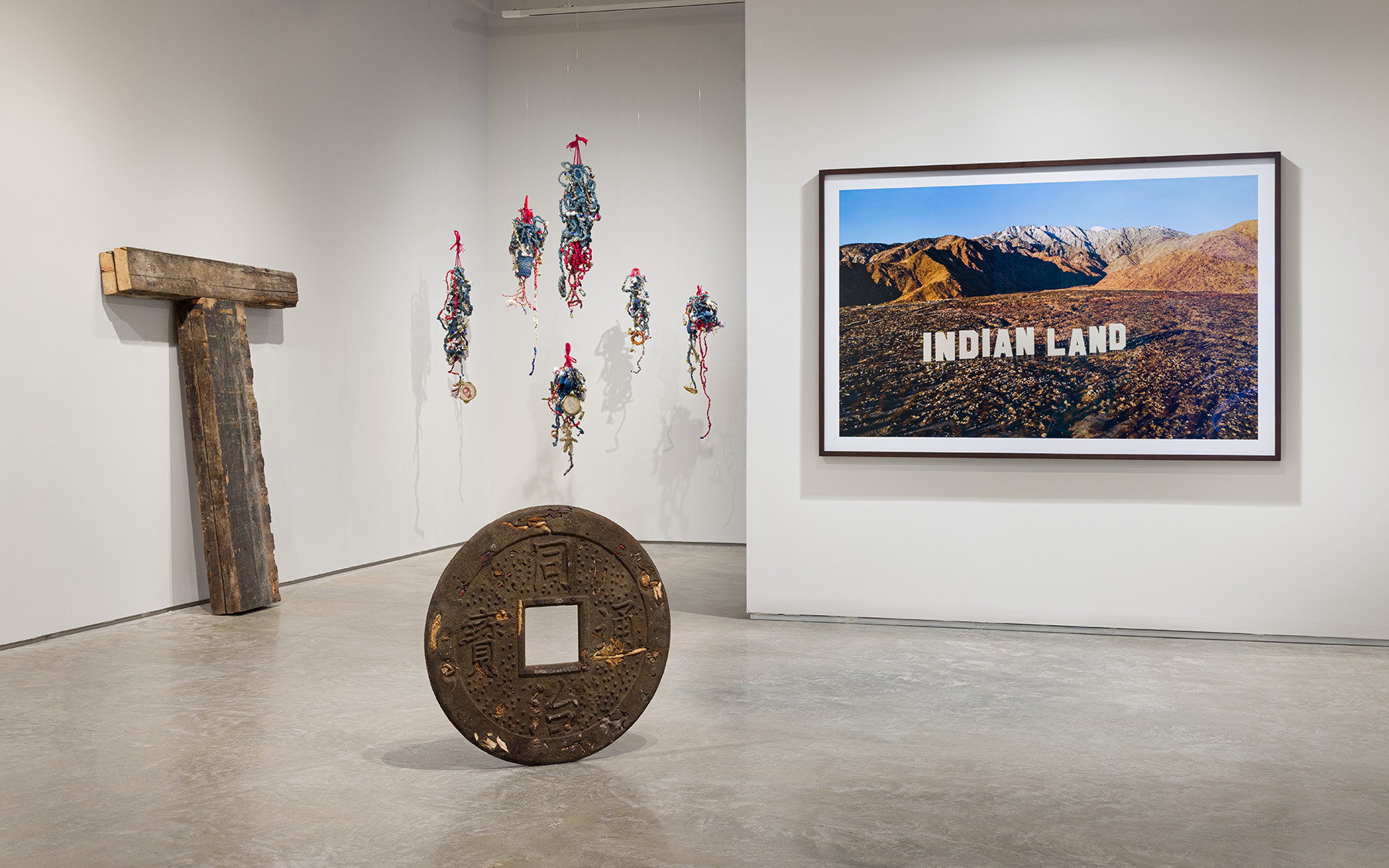 An Incomplete Haunting, Installation view. From left to right: Kris Grey, Capital T, 2025. Salvaged wooden joists from Stonewall National Monument Visitor Center, 75 x 60 x 6 inches. Courtesy of the artist. Elvira Clayton, Indigo Bess and her Children, 436 project, 2024. Six sculptures: indigo-dyed osnaburg, cotton, wire, jute, sequin, mini glass bottles, cotton bolls, shells, twigs, screen-printed archival text and photographs, embroidery hoops, acrylic, handwoven, and crochet. Bess: 37 x 12 x 7 inches; Sam: 46 x 12 x 5 inches; Elizabeth: 37 x 7 x 5 inches; Sena: 29 x 10 x 7 inches; Affa: 31 x 7 x 4 inches; Hannah: 37 x 10 x 6 inches. Courtesy of the artist. Kenneth Tam, Why do you abuse me, 2022. Epoxy resin, dirt, sand, dried mushrooms, dried seaweed, dried bamboo shoots, dried jujube, dried goji berries, preserved apricots, sunﬂower seeds, dried roots, dried sweet potato, steel, 30 x 30 x 1 1/2 inches. Courtesy of the artist and Commonwealth and Council. Nicholas Galanin, Never Forget, 2021. C-print mounted on Dibond, walnut frame, 51 3/4 x 78 3/4 inches. Courtesy the artist and Peter Blum Gallery, New York. Photo by Etienne Frossard.