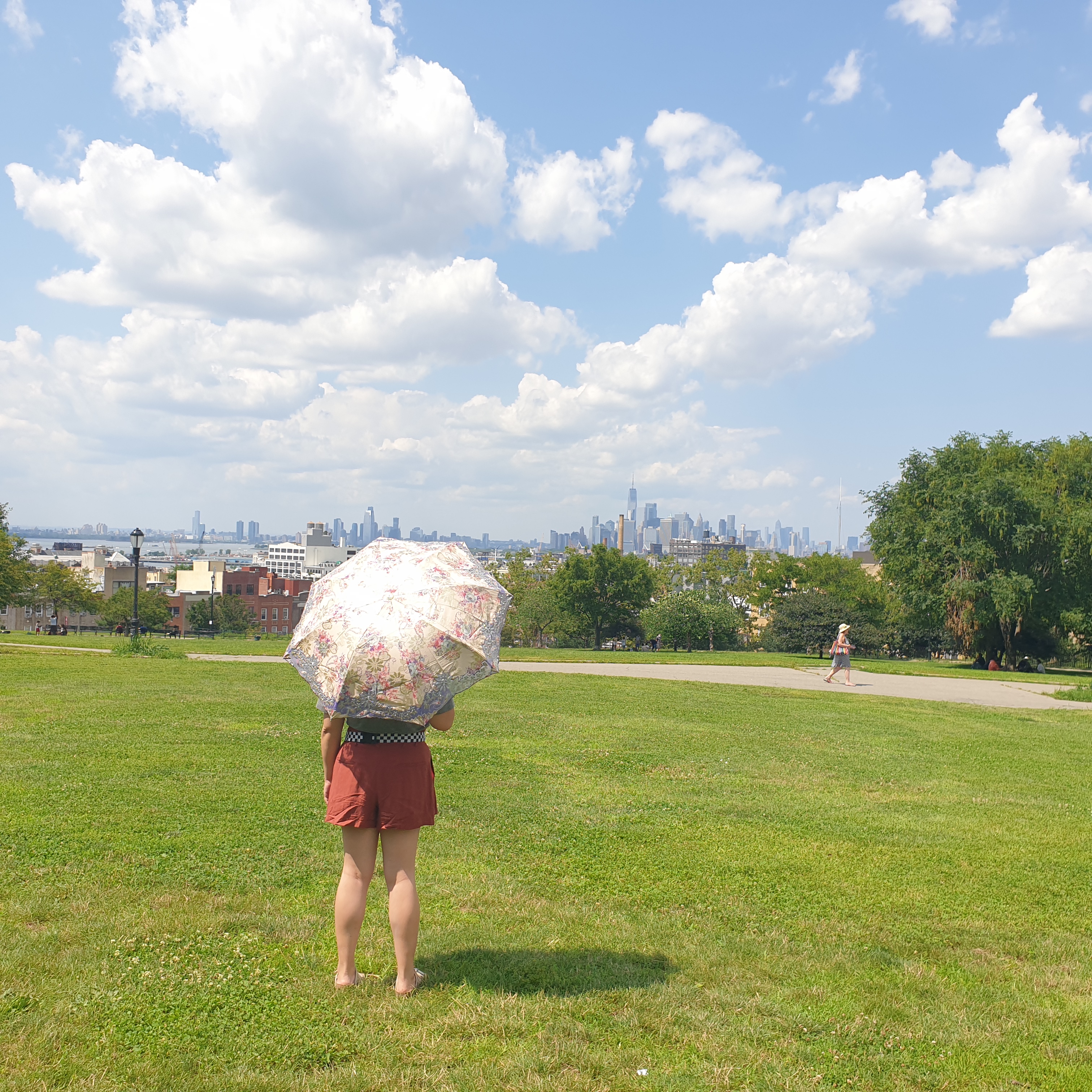 A woman stands on green grass holding a purple and gold parasol. The skyline of manhattan can be seen in the background under a bright sky with large white clouds. A person walks in the middle of the picture through the asphalt path in Sunset Park.