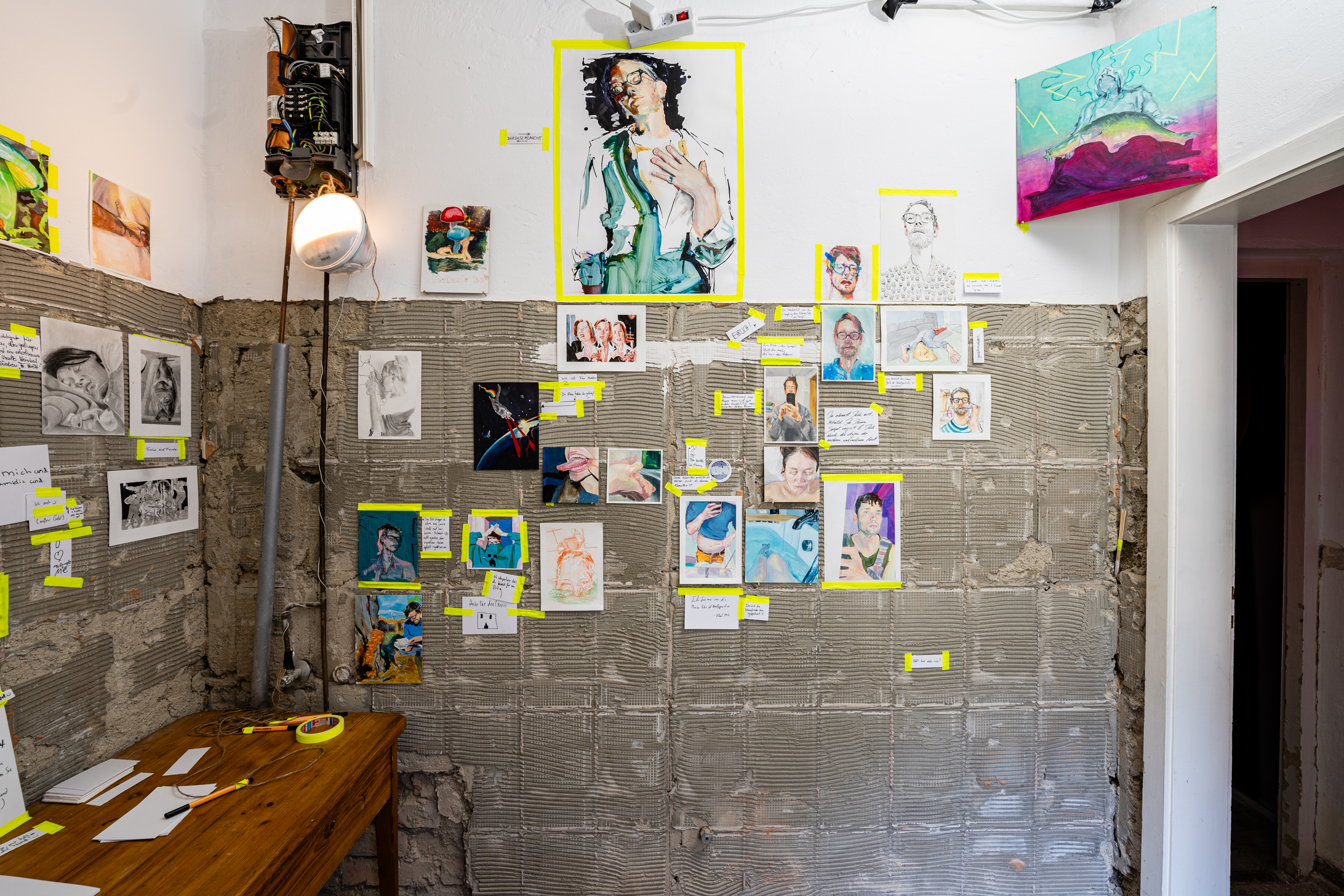 Bild 1 zu Kunst kommt von Kompostieren: Wand rechts. An der Wand h&auml;ngen verschieden gro&szlig;e und kleine Malereien, die mit gelbem Klebeband befestigt wurden. Die Wand ist zum Gro&szlig;teil unverputzt, sodass das Gem&auml;uer zu sehen ist.