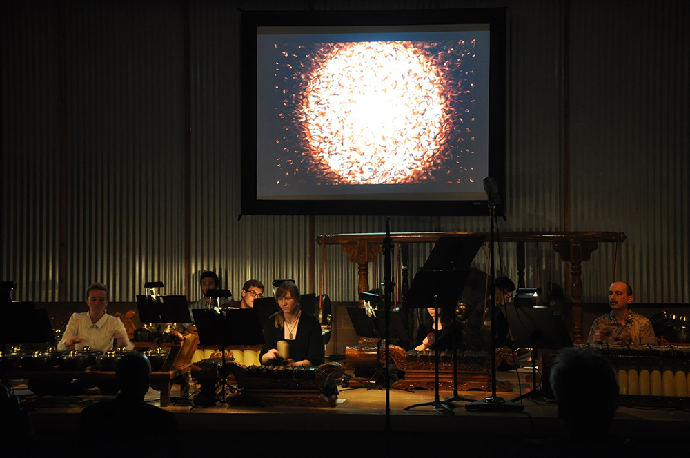 Musicians of the HMC American Gamelan perform on a dark stage. A video art is displayed on screen in the back and above.