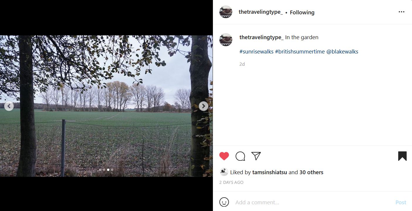 Instagram post by @thetravelingtype_ of A field framed by trees with leaves that are turning colour. There is a green field extending to teh foreground, with another line of trees and an overcast sky visible beyond. The text reads: "thetravelingtype_'s profile picture thetravelingtype_ In the garden  #sunrisewalks #britishsummertime @blakewalks"