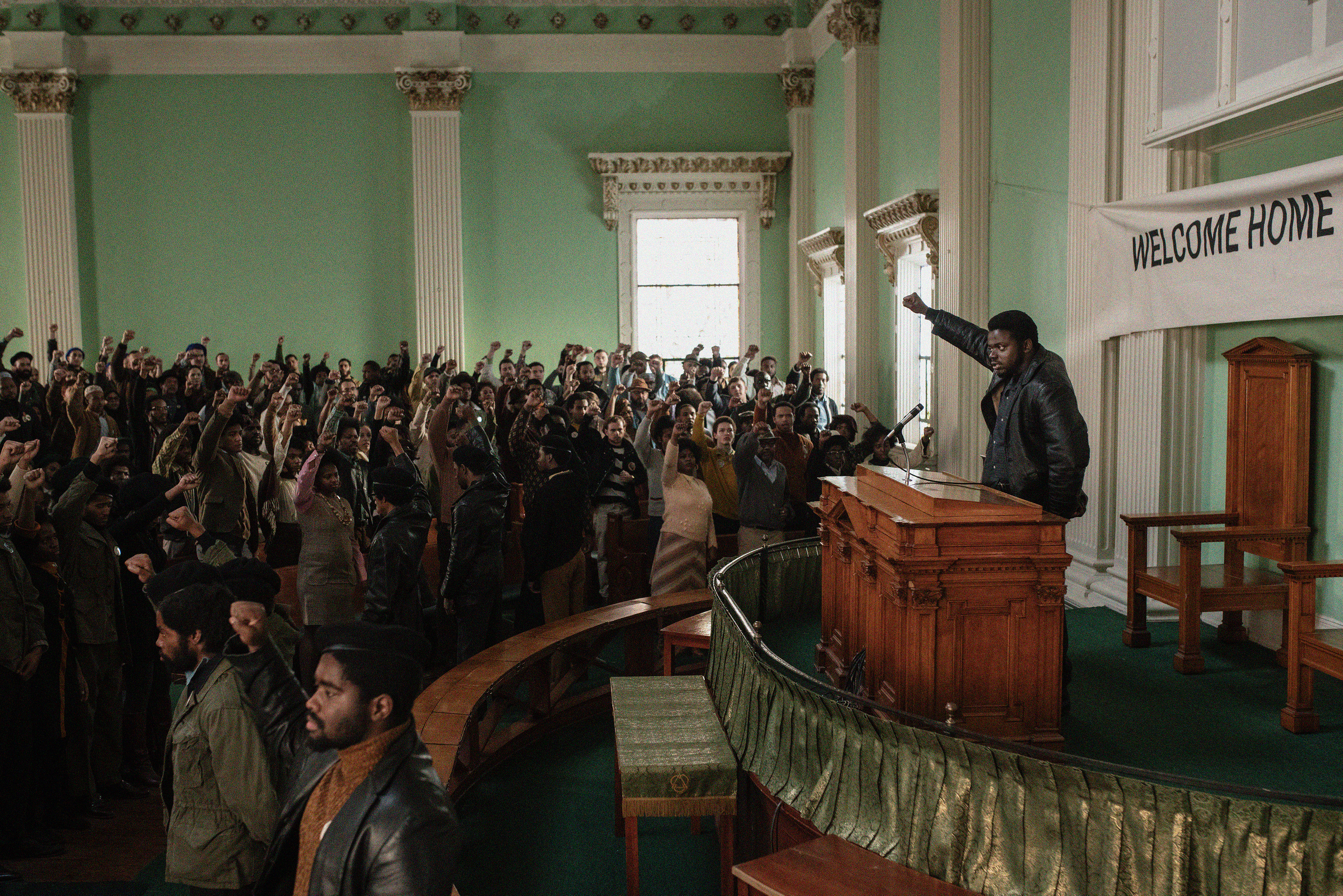 Image Description: Still from "Judas and the Black Messiah" picturing a large meeting space, resembling the inside of a church or community meeting hall. The large audience is predominantly made up of Black people who stand, raising their fists in the sky and looking towards a stage, where a young man faces them at a podium and holds his fist towards the sky as well.