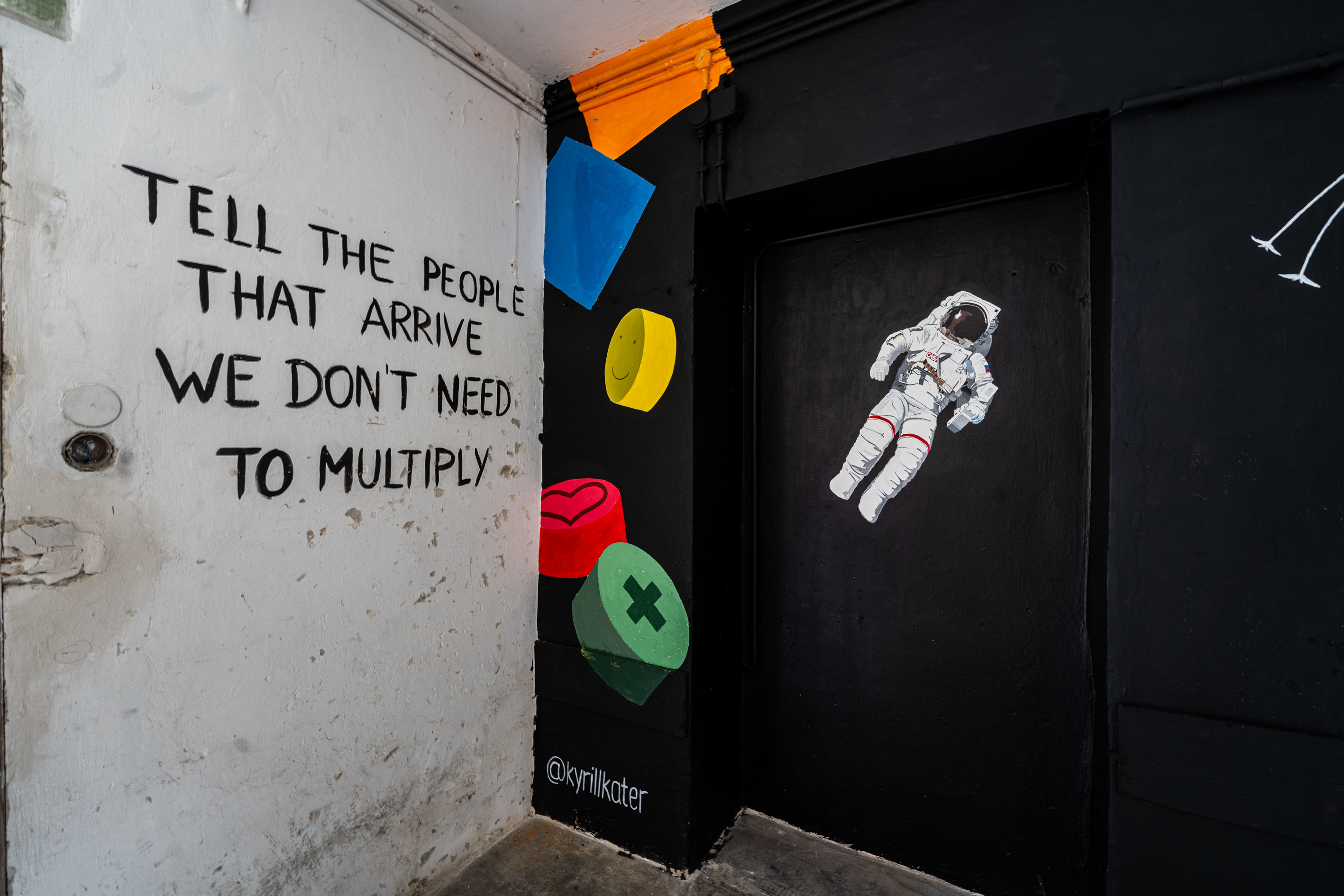 Bild 2 zu Mein Leben in vier W&auml;nden: Die linke Wand des Vorraumes. Diese ist schwarz. In der Einbuchtung der Wand ist ein realistisch gemalter Astronaut mit wei&szlig;em Anzug erkennbar. Links davon bunte geometrische Formen. An der Wand der Eingangst&uuml;r steht in schwarzer Handschrift: "tell the people that arrive we don`t need to multiply".