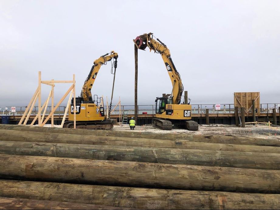 Construction photo showing two excavators installing large timber piles into the ground for the elevated foundation of the Brigantine Marina Paddle Club.