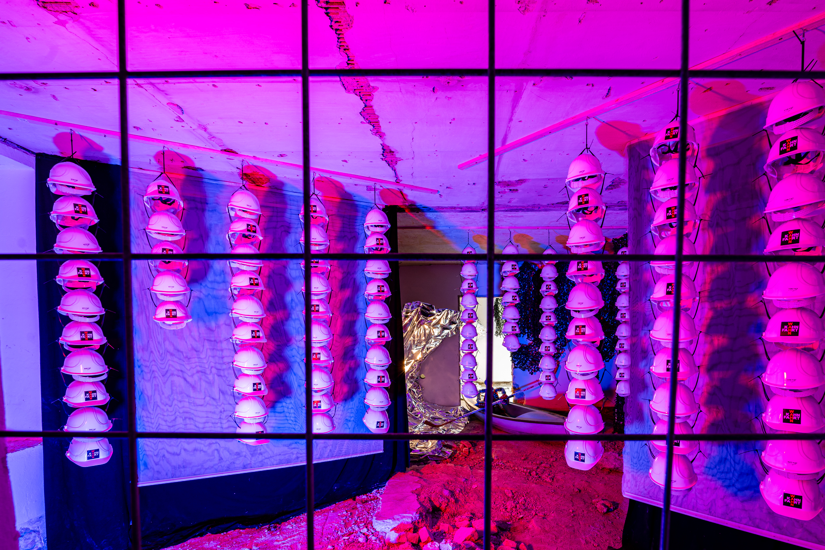 Bild 1 zu Wahnfahrt:  Durch ein Absperrgitter kann lediglich von einer Stelle in den gro&szlig;en Raum geblickt werden. Hierbei kommen verschiedene optische Effekte zum Einsatz.  Die Installation ist in blaues Licht getaucht, das kurz durch rote Lichtblitze gebrochen wird. Hierdurch entsteht der Eindruck, als w&uuml;rde der Schriftzug &bdquo;Wahn&ldquo; der Helme rot aufleuchten.  Auf den doppelt aufgeh&auml;ngten Vorh&auml;ngen links und rechts im Vordergrund ist der sog. Moir&eacute;-Effekt zu sehen, der auf eine kr&auml;uselnde Wasseroberfl&auml;che anspielt.  Durch diese Begrenzung der Sichtachsen in Kombination mit Theaterlaub sind Fenster, T&uuml;ren, Unterz&uuml;ge und Raumst&uuml;tze des urspr&uuml;nglichen vorgefundenen Raumes kaschiert. Im hinteren Teil des Raumes liegt auf dem Boden, der teils mit Schutt bedeckt ist, ein Originalboot der Kahnfahrt. Darin sitzt eine Figur in Eishockey-Montur, ein Querverweis auf die Probleme beim Umbau des Curt-Frenzel-Stadions.