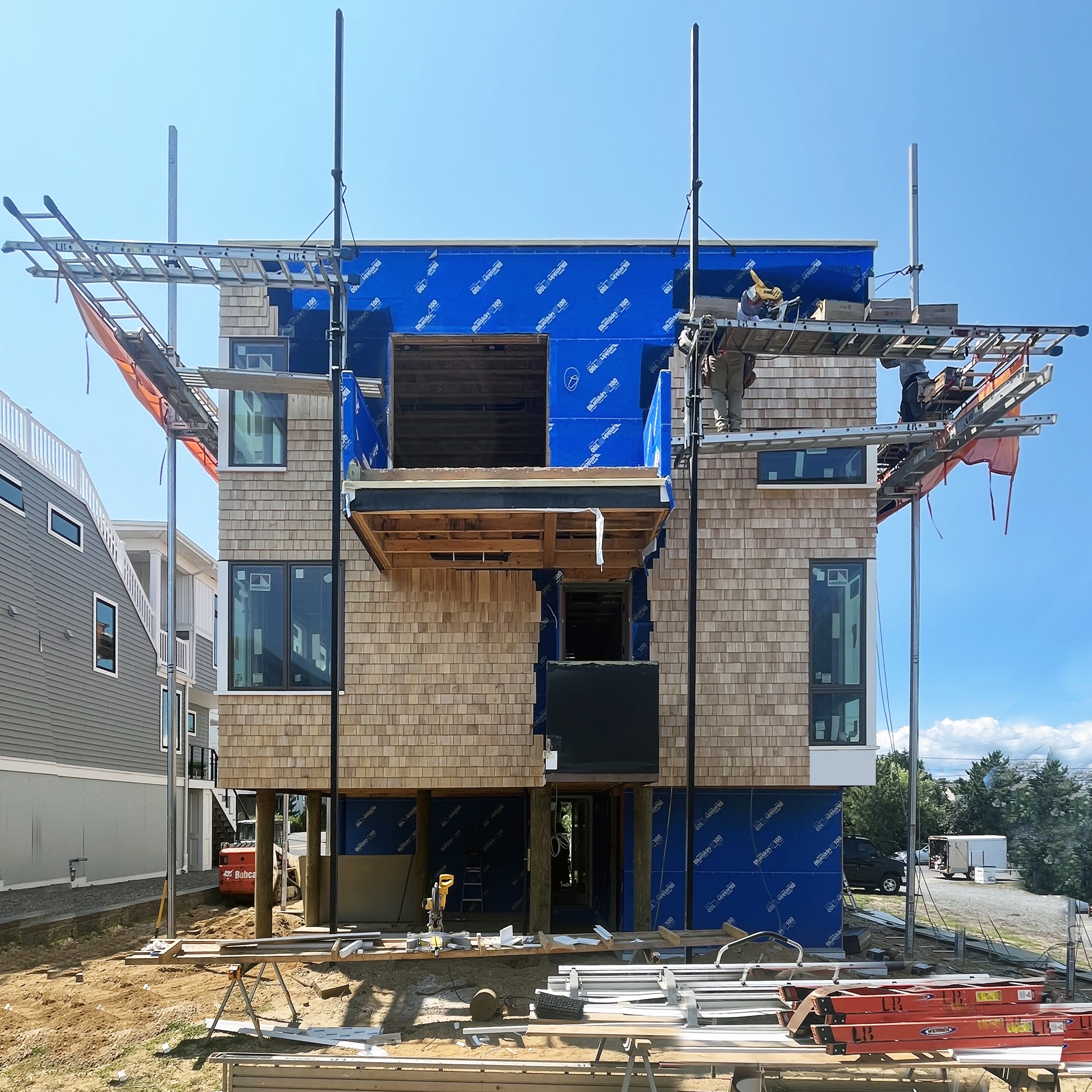 Construction photo of The Gap House exterior, showing the installation of the cedar shingle cladding and blue weather barrier with scaffolding in place.
