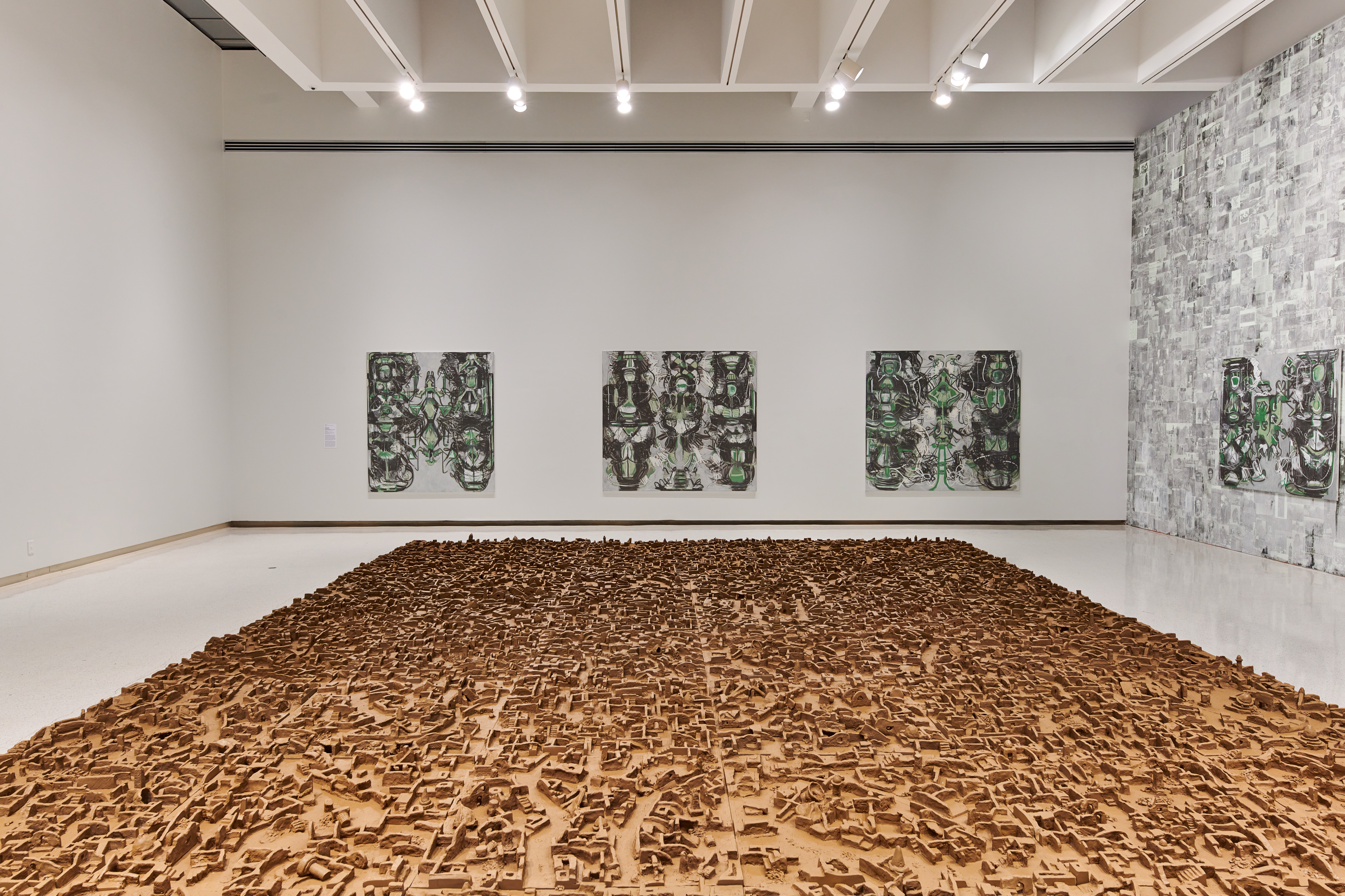 A white wall gallery space that has a floor installation taking up nearly the entire room of a sand colored, maze-like shallow relief, that resembles a large city as if seen from far above. Four larger paintings line the walls behind.
