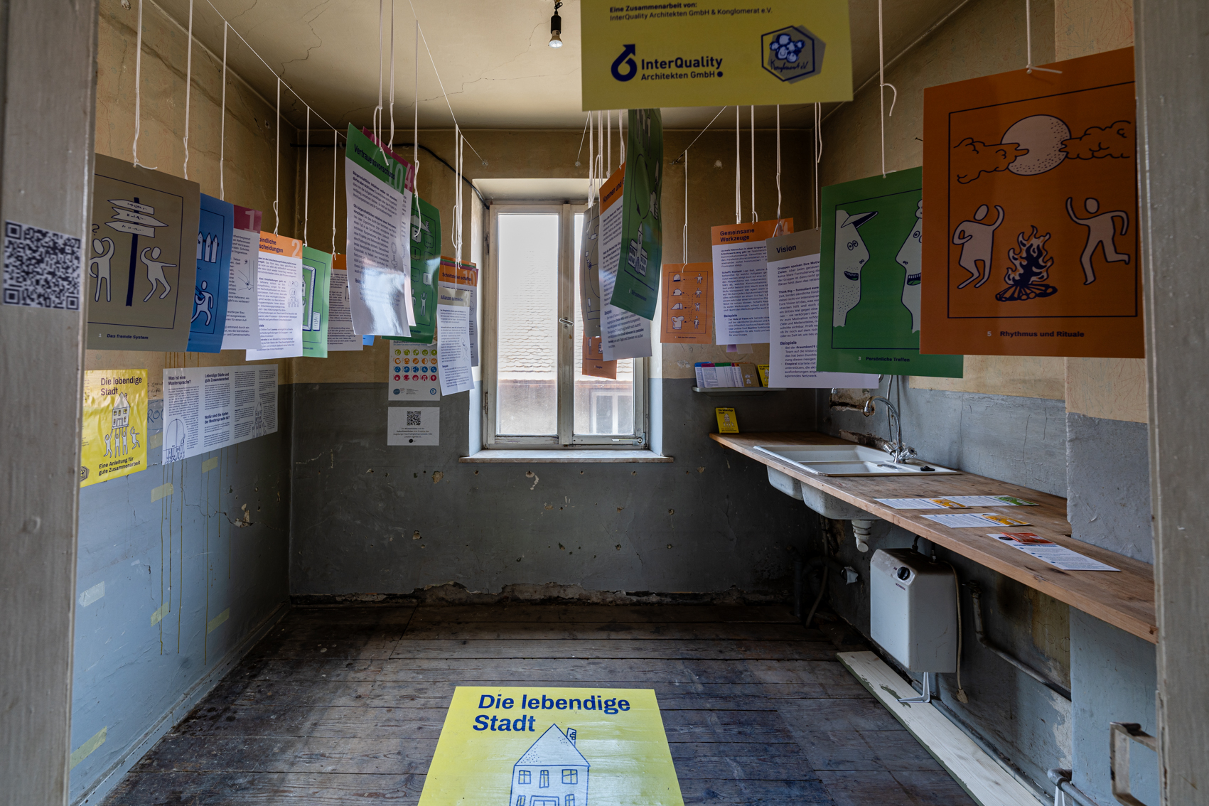 Bild 1 zu Musterk&uuml;che:  Blick durch die T&uuml;r in den Raum. Gegen&uuml;ber mittig ein Fenster. An der rechten Wandseite befindet sich ein Holzbrett mit eingebauter Sp&uuml;le. Von der Decke h&auml;ngen an F&auml;den  Karten des Sets "Die lebendige Stadt".