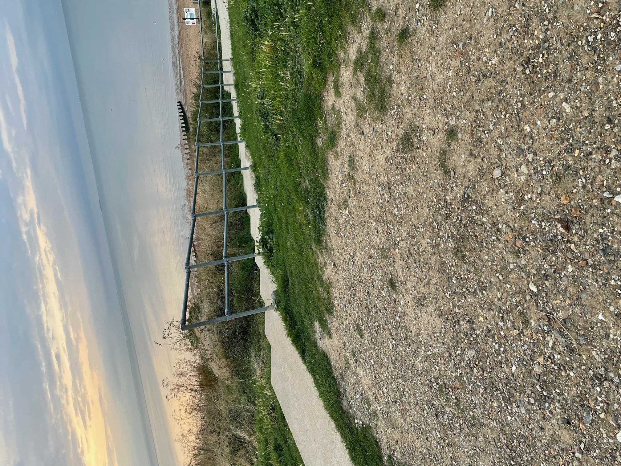 Stairs lead down to the beach with gravel and grass on either side. The ocean can be seen in the distance, as well as a sliver of beach.