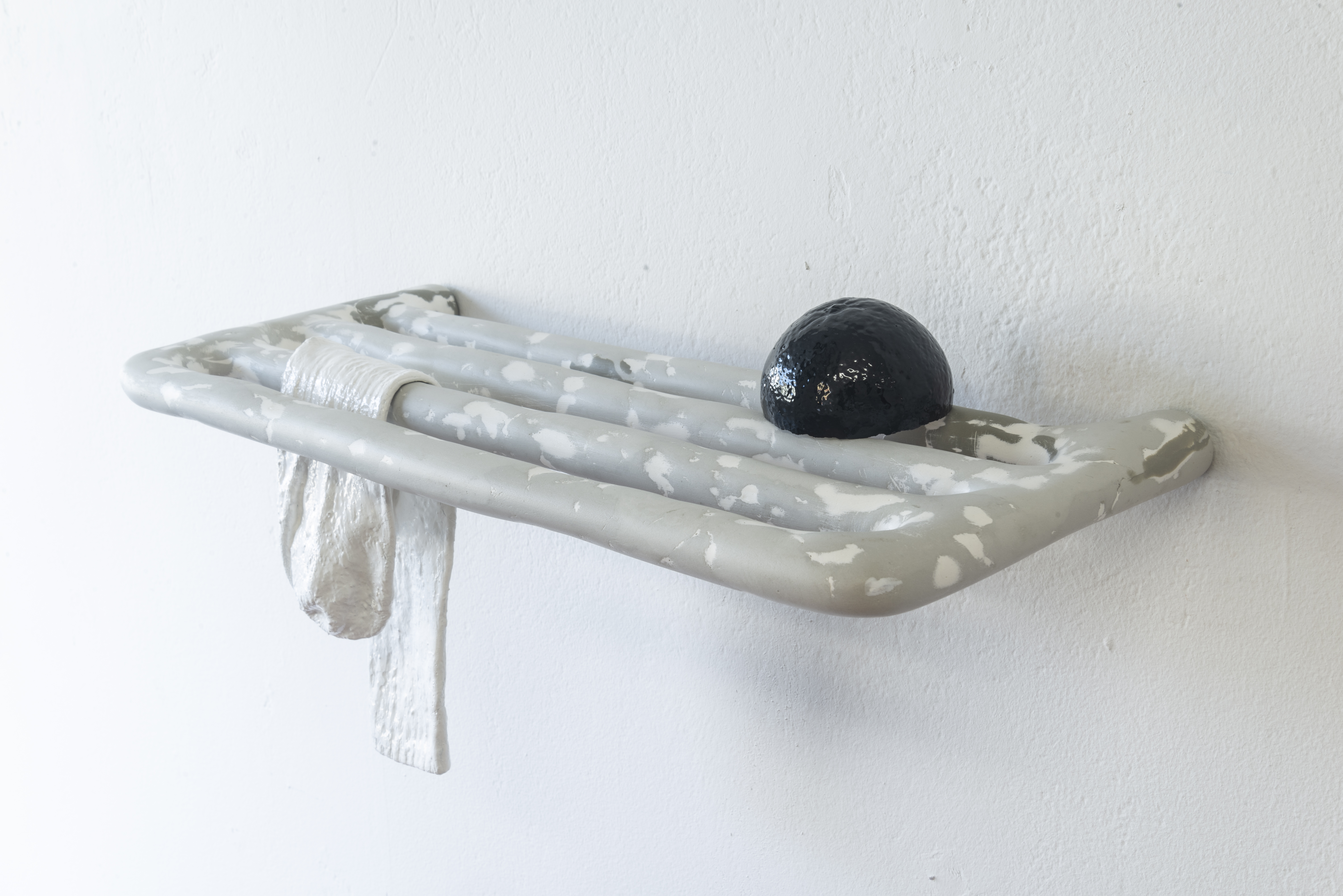 Sculpture of a grey, clearly handmade, clothing rack that protrudes impossibly out of the wall with another sock and ambiguous black orbital mass slouching into the slats.