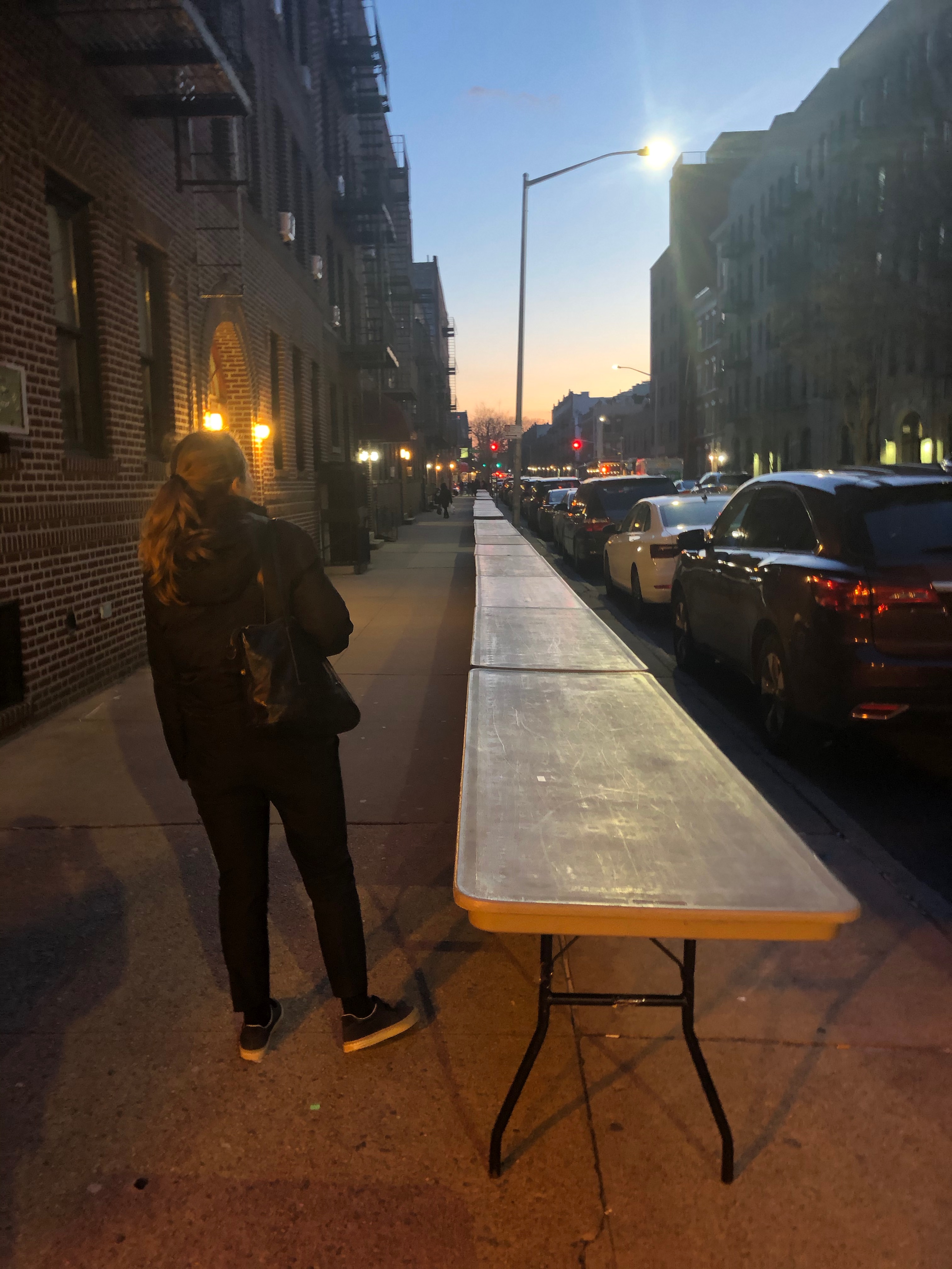 A set of empty foldable tables are set up along the sidewalk. A woman stands next to them with her back to the camera.