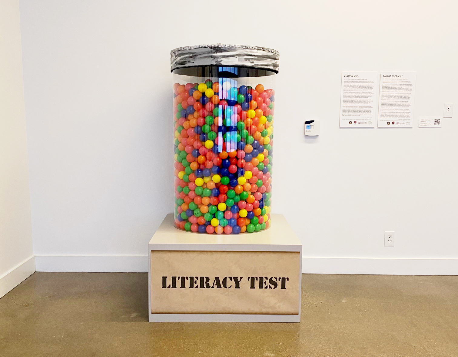 A person-sized glass cylinder with a shiny steel cap filled with thousands of plastic balls, suggested to be oversized gum balls, all sit atop a pedestal that read “literacy test.”