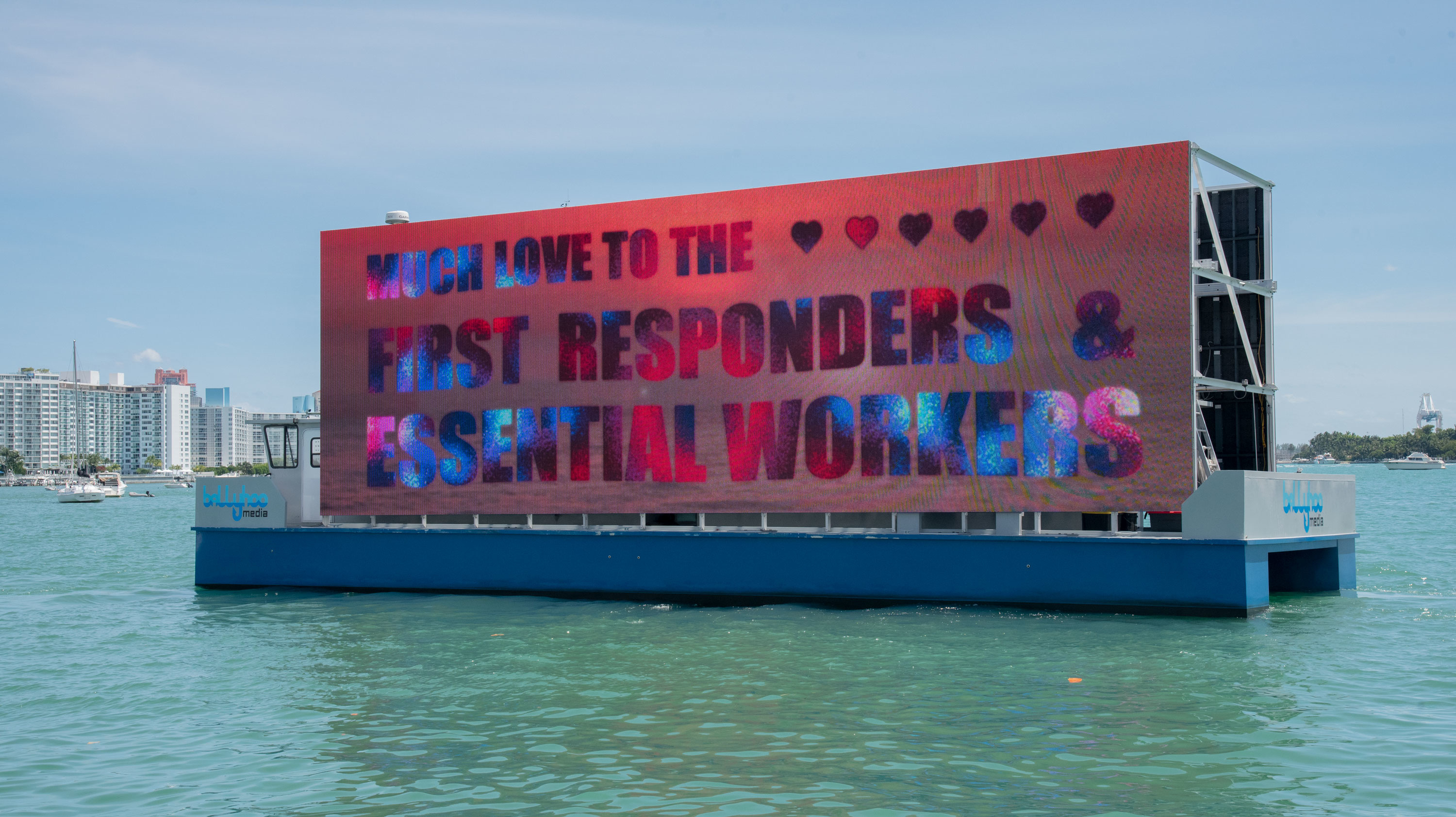 Displayed on a large screen that is being exhibited by boat, Carlos Betancourt’s 2020 video installation projects the text: “Much Love to the First Responders & Essential Workers”, in large block letters accompanied by hearts, on an ocean sunset background.