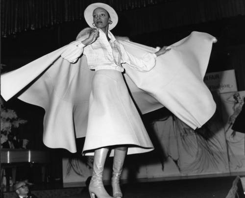 Image: A black-and-white photo of an Ebony Fashion Fair model posing on a runway in a flowing and flared out light-colored cape and skirt, with a button up blouse and hat, wearing leather boots, c. 1974. African American Photographs Collection, Tom and Ethel Bradley Center, California State University Northridge. Photographer: Guy Crowder.