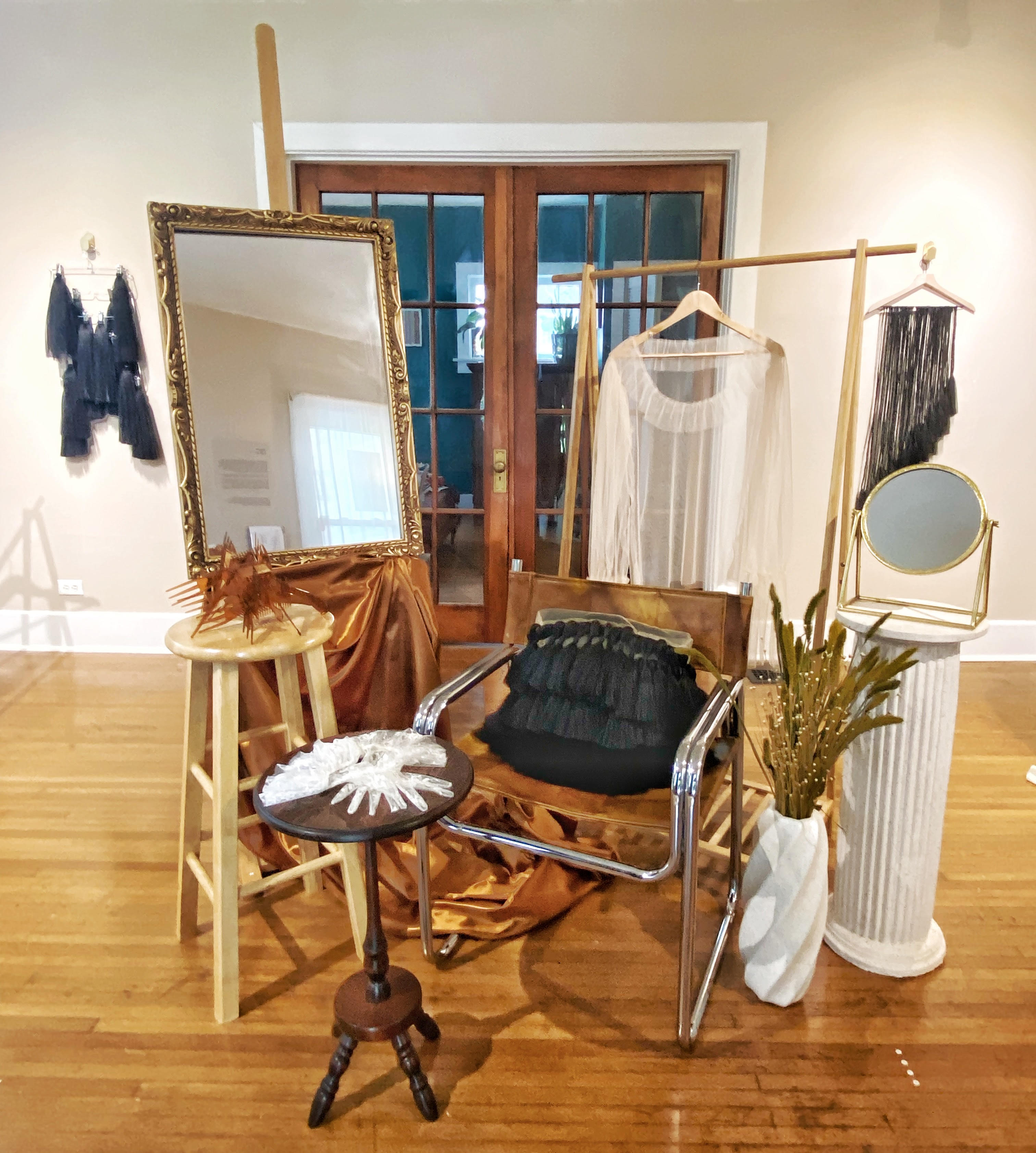 A scene of carious sculptures inn an empty hardwood floor living room, predominantly made from found domestic materials, here all together, resembling a kind of salon or barbershop, with a central chair, various mirrors and size tables, a hip high Romanesque column, a white shirt on a clothes hanger, and so on.