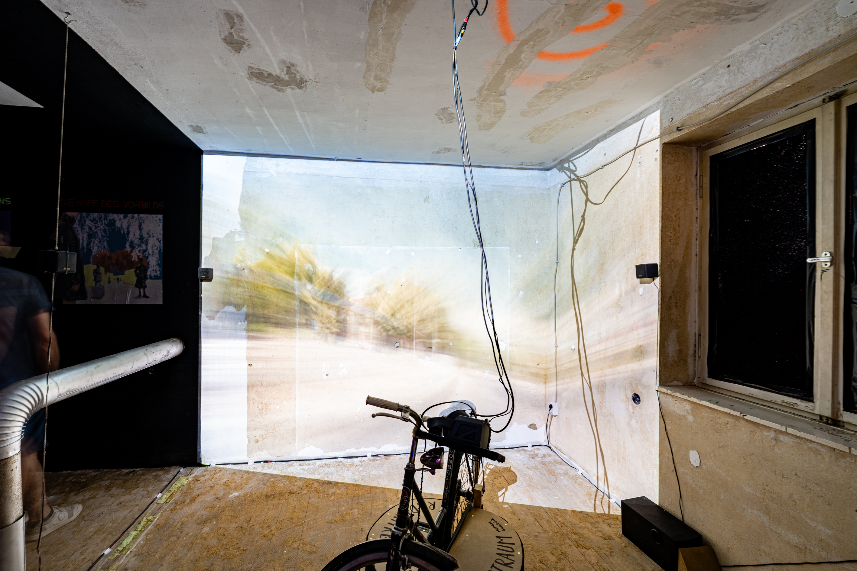 Bild 1 Radfahren: Ein Fahrrad steht in der Mitte des Raumes. Dahinter wirft ein Beamer die vorbeiziehenden Stra&szlig;en Augsburgs auf die Wand. 