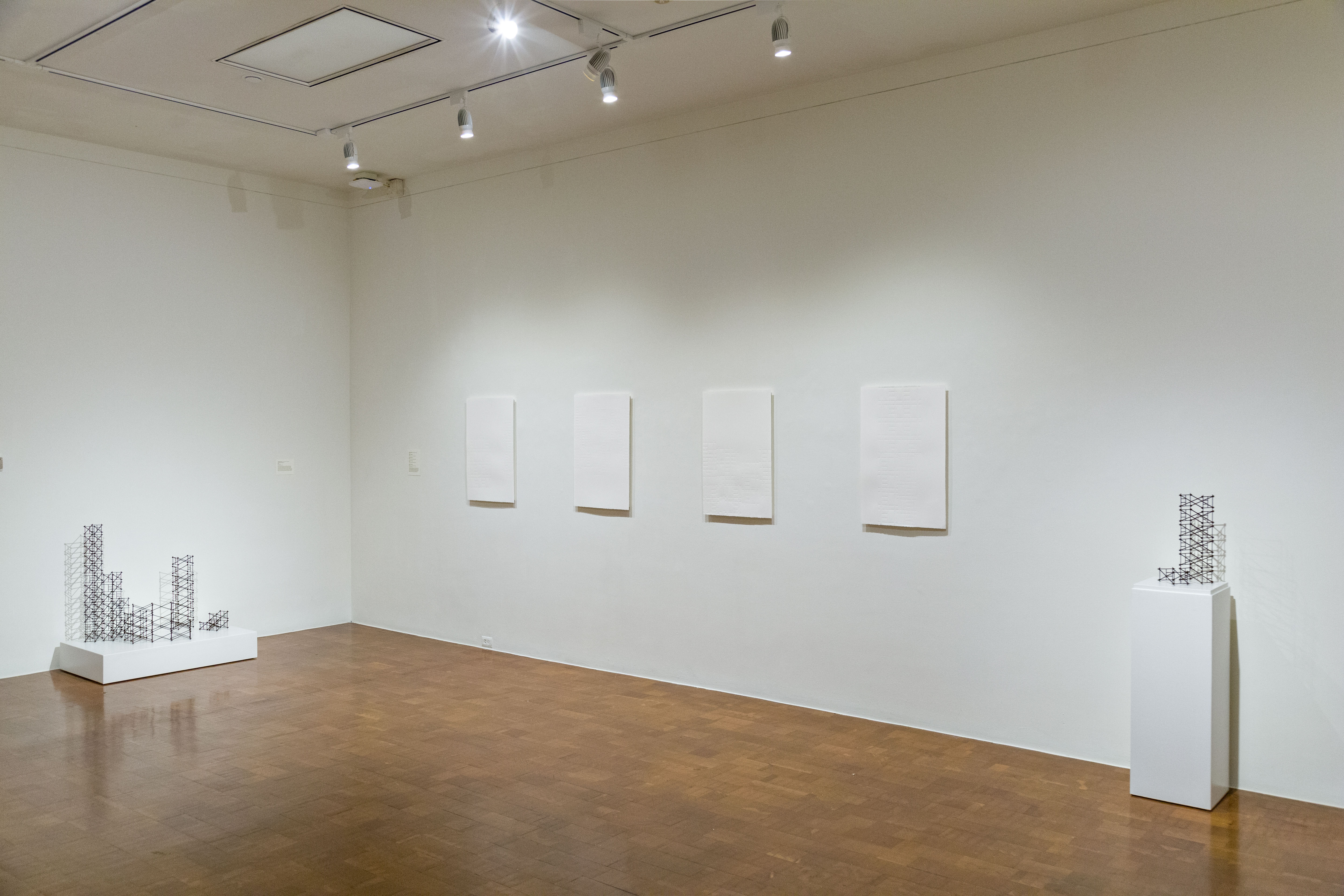 View of a sparse, white wall museum space with shiny wood floors and track lighting overhead. On the far left and right are pedestals with small wireframe sculptures that loosely resemble buildings, and in between on the wall is four, seemingly blank canvases, all hung in a row.