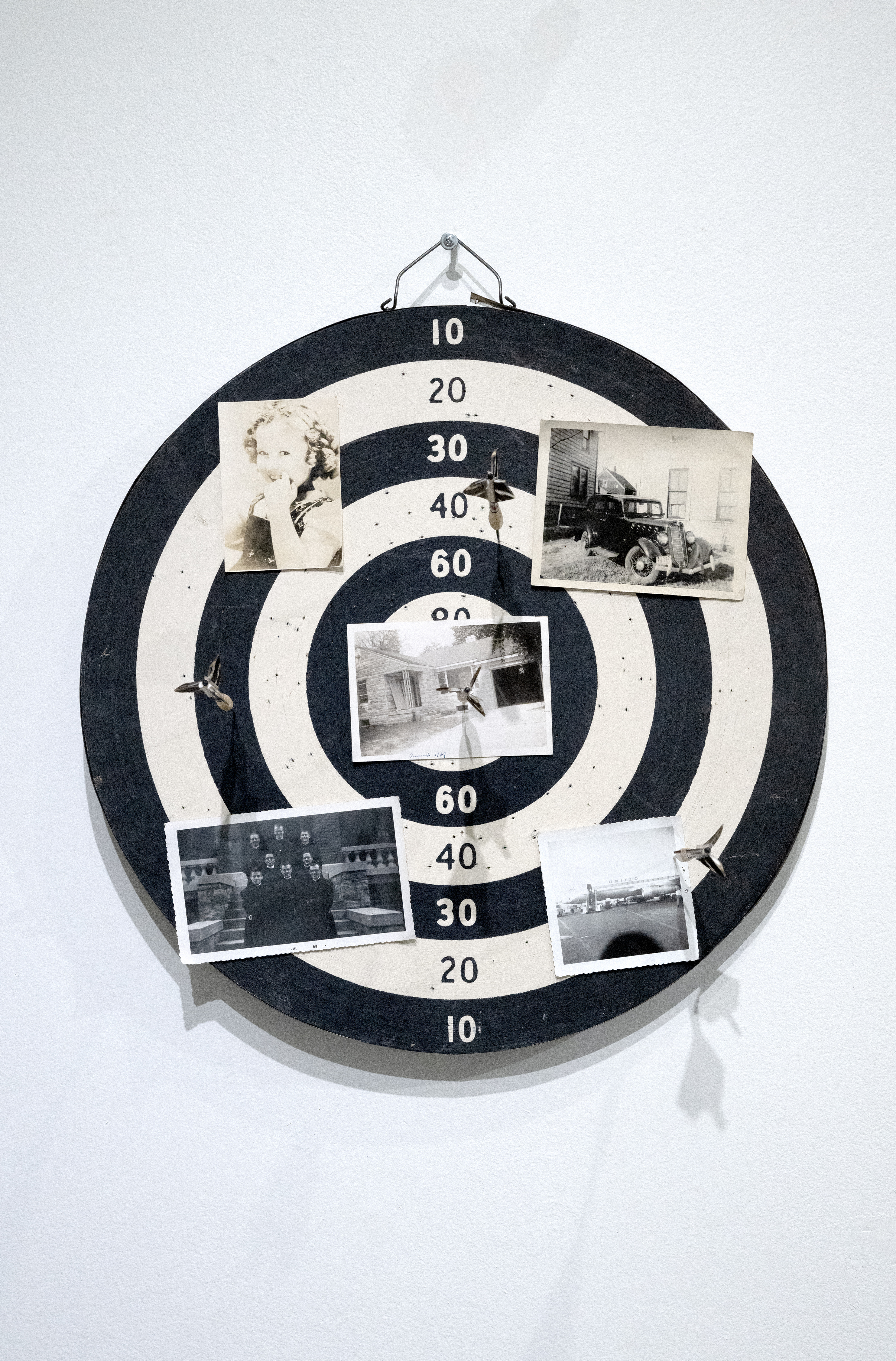 Assimilation, Campbell Hall Studio, Studio City, California (2024). Through a conceptual lens, the arrangement of darts and archival photographs on a dart board explores the internal landscape of forced assimilation within the American dream. 